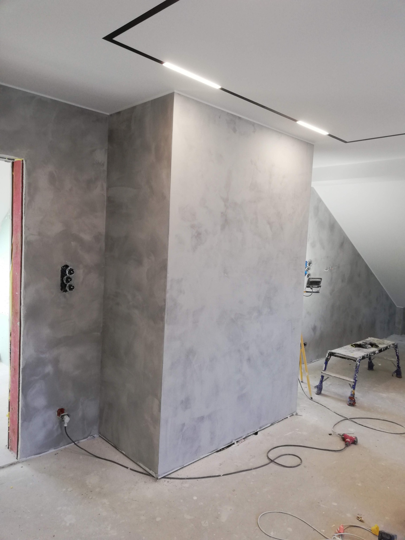 Ściana w pokoju pokryta szarą, dekoracyjną gładzią w stylu weneckim, z widocznymi gniazdkami elektrycznymi i czarną listwą oświetleniową sufitową; na podłodze narzędzia i drabina malarska.