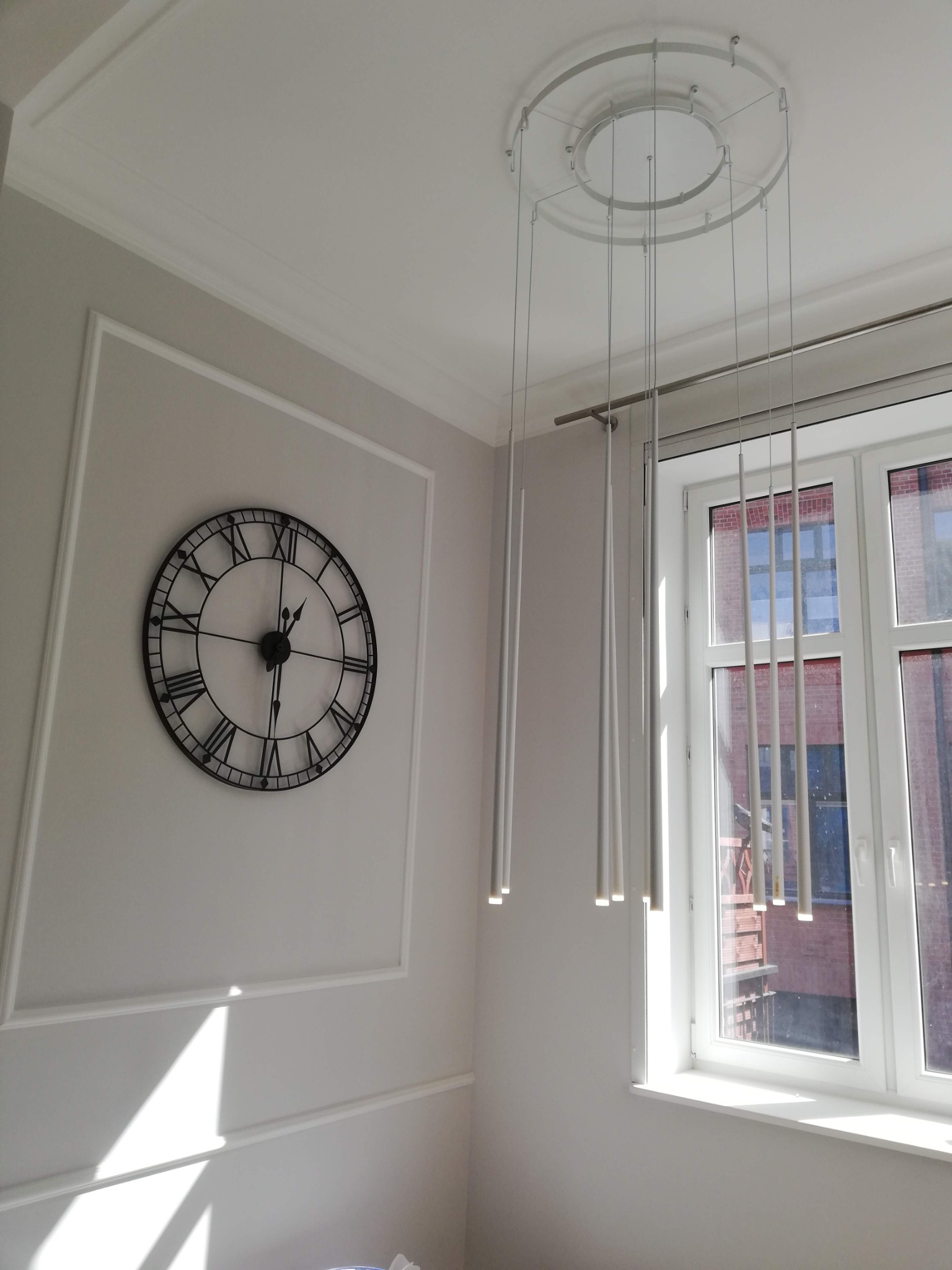 Salon w jasnych barwach z dekoracyjnym zegarem na ścianie z listwami, nowoczesną lampą wiszącą z cylindrycznymi elementami oraz oknem z widokiem na budynek z czerwonej cegły.