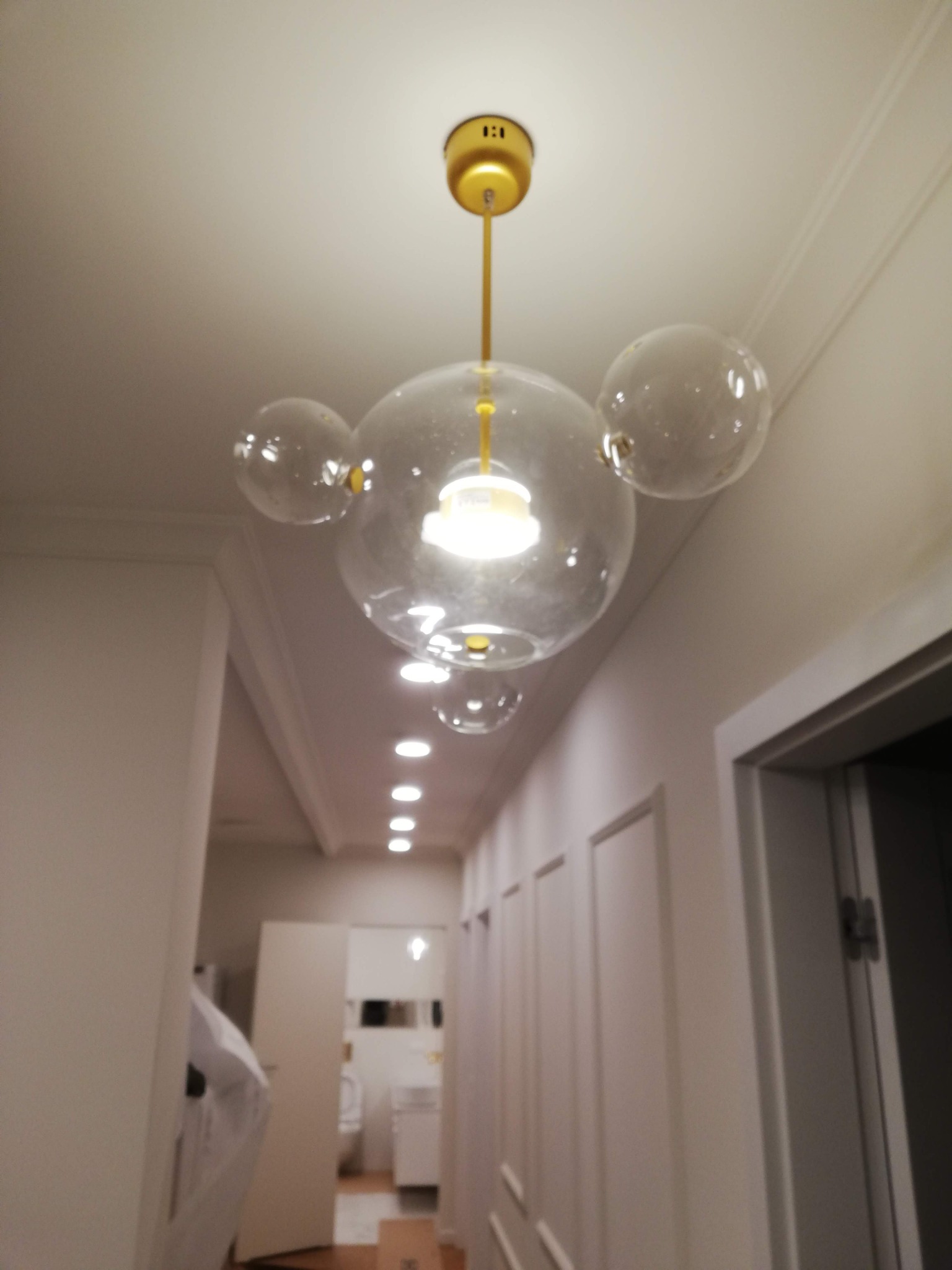 Szklany żyrandol w kształcie kuli z trzema mniejszymi kulami, zawieszony na złotym pręcie w białym korytarzu z widokiem na łazienkę w tle.