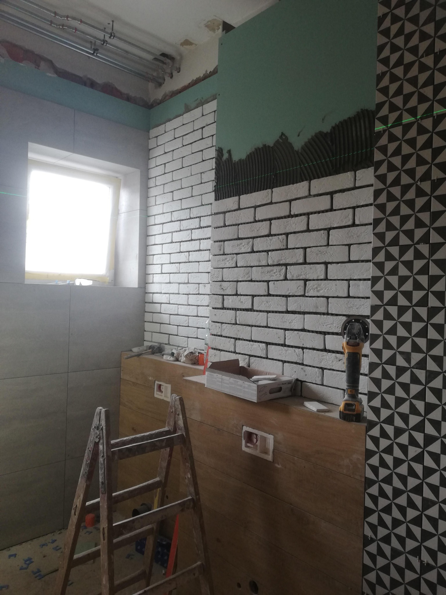 Wnętrze łazienki w trakcie wykańczania: biała cegiełka na ścianie, płytki z geometrycznym wzorem, drewniana zabudowa pod umywalkę, drabina malarska i szlifierka kątowa na blacie.