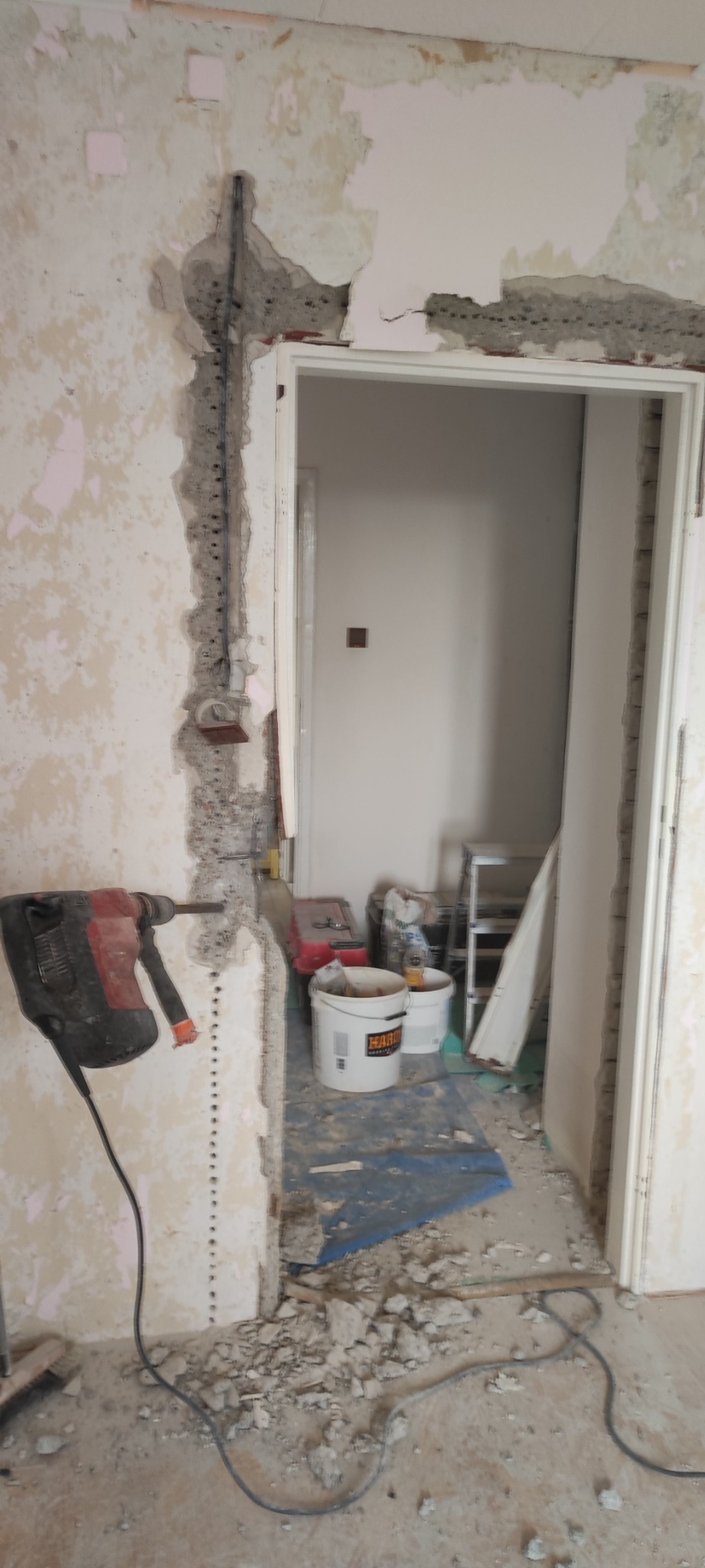 Wnętrze w trakcie prac remontowych: ściana z widocznymi śladami po kuciu, instalacja elektryczna, wiertarka udarowa, wiaderka z zaprawą i gruz na podłodze przy framudze drzwi.