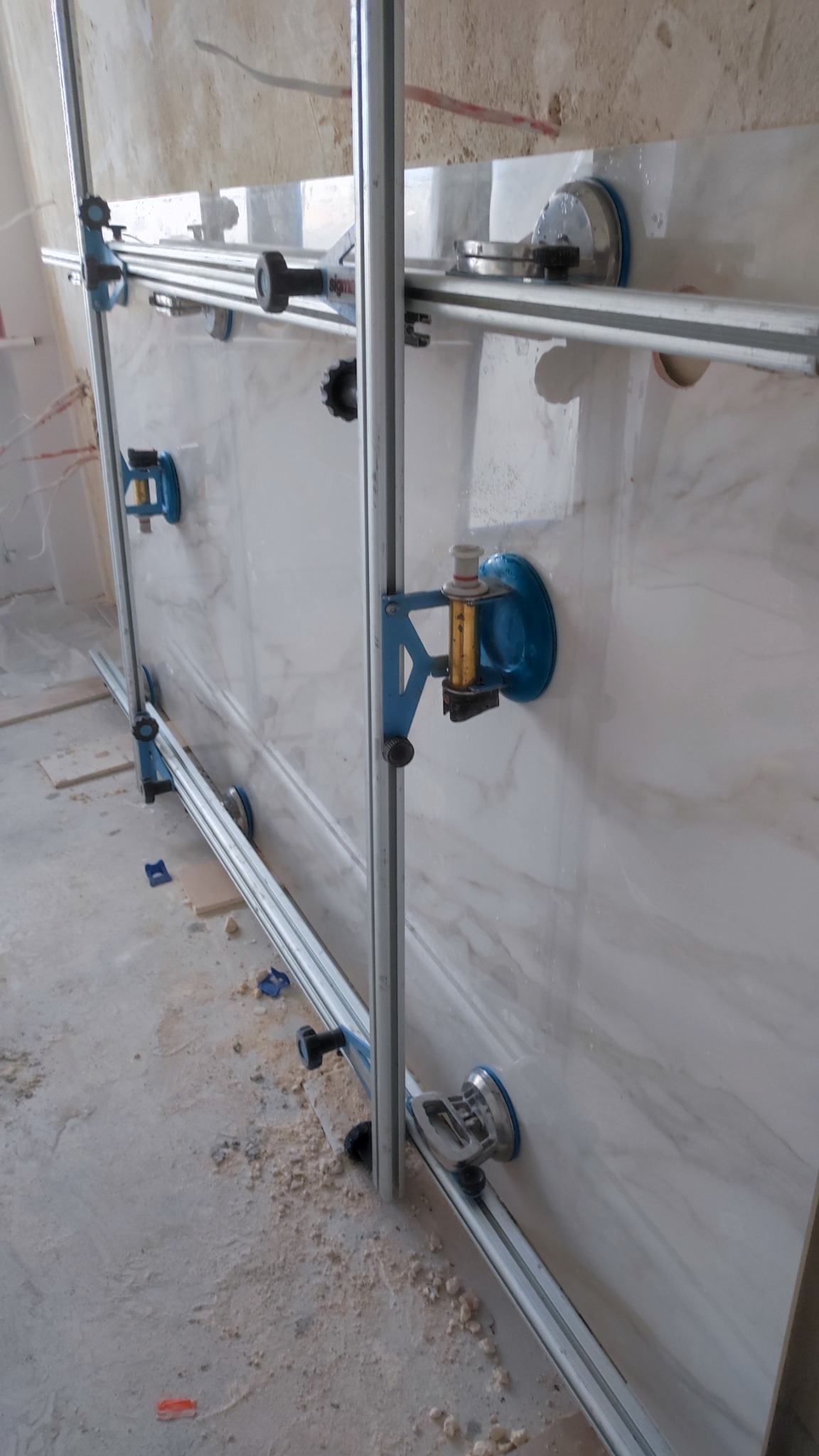 Montaż wielkoformatowej płyty imitującej marmur za pomocą specjalistycznego systemu szyn i przyssawek, widoczne narzędzia i drobne zabrudzenia na podłodze.