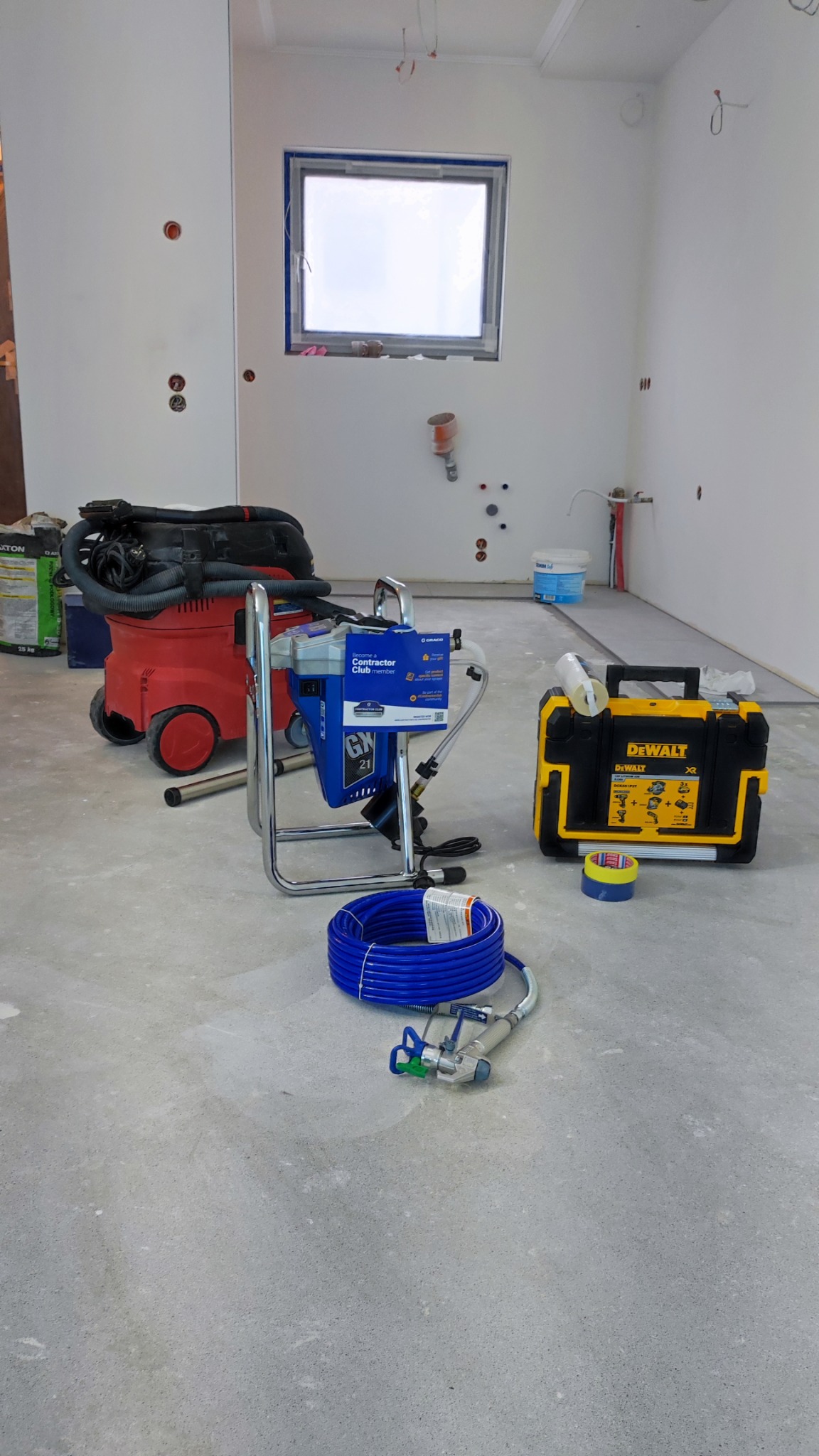Pokój w trakcie remontu z widocznym agregatem malarskim Graco, odkurzaczem przemysłowym na kółkach, niebieskim wężem, pistoletem natryskowym i skrzynką narzędziową DeWalt, ściany przygotowane...
