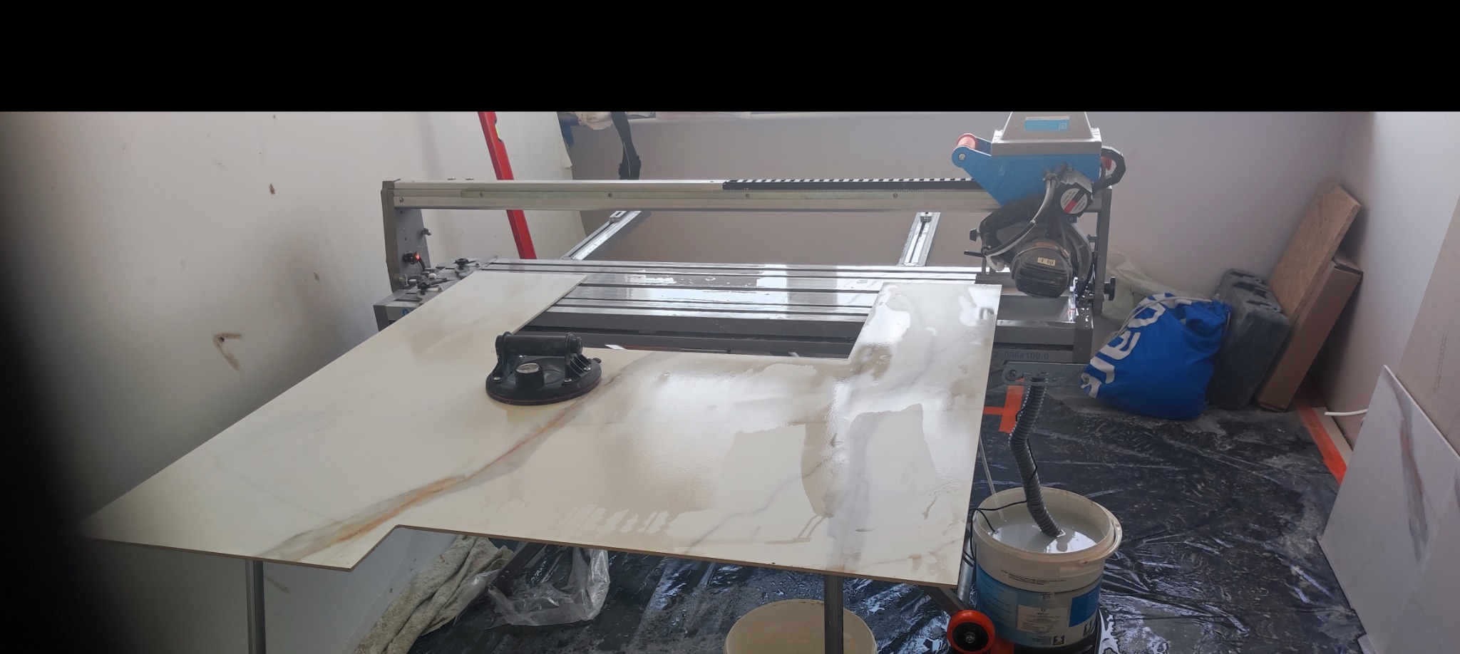 Płytka ceramiczna z marmurkowym wzorem, przycięta z otworem, leży na stole roboczym z przyssawką, obok stoi profesjonalna przecinarka do płytek, na podłodze folia ochronna.