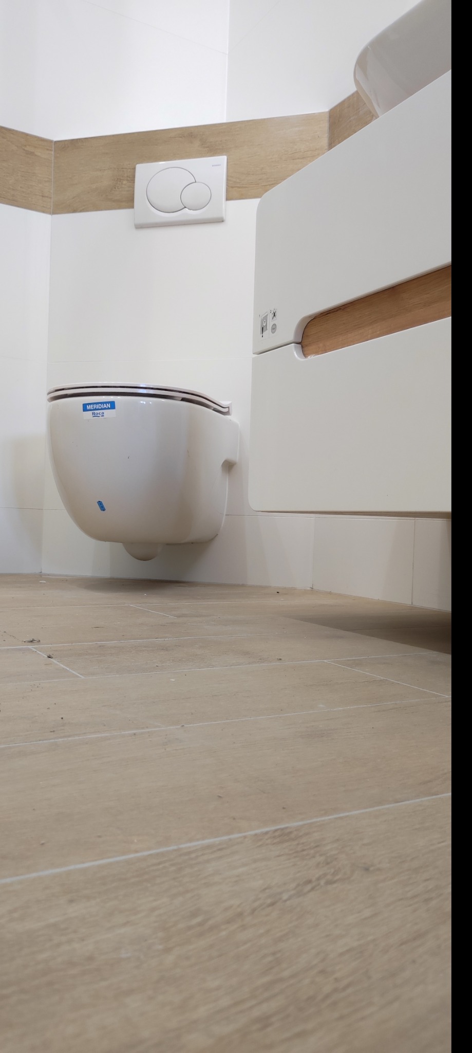 Wisząca toaleta Roca Meridian z podtynkową spłuczką Geberit i szafką z drewnianym akcentem w wykończonej łazience z beżowymi płytkami podłogowymi.