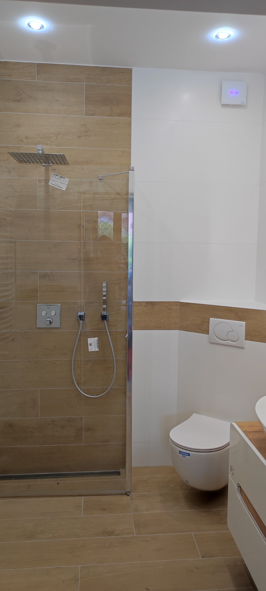 Nowoczesna łazienka z kabiną prysznicową typu walk-in, wykończona płytkami imitującymi drewno i białymi panelami ściennymi, z widoczną toaletą i elementami armatury.