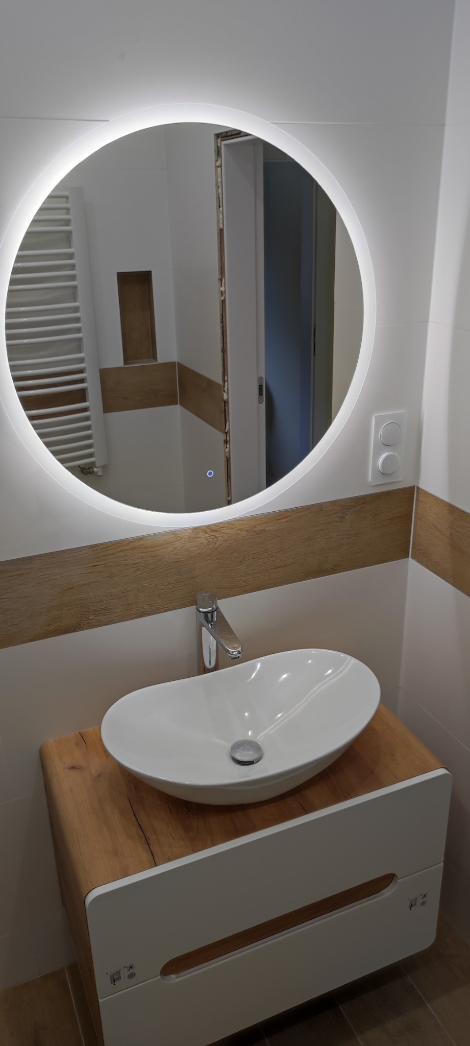 Owalna umywalka nablatowa z chromowaną baterią na szafce z drewnianym blatem, okrągłe lustro z podświetleniem LED, fragment wykończonej łazienki z płytkami imitującymi drewno.