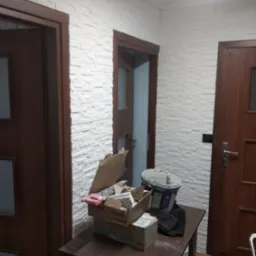 Wnętrze korytarza z trzema drewnianymi drzwiami i ścianą pokrytą dekoracyjną białą cegłą, na pierwszym planie stół z narzędziami i pojemnikiem z zaprawą.