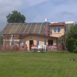 Dom w trakcie remontu dachu, widoczne krokwie i membrana dachowa, częściowo położona dachówka, okna i drzwi przygotowane do montażu na trawniku.