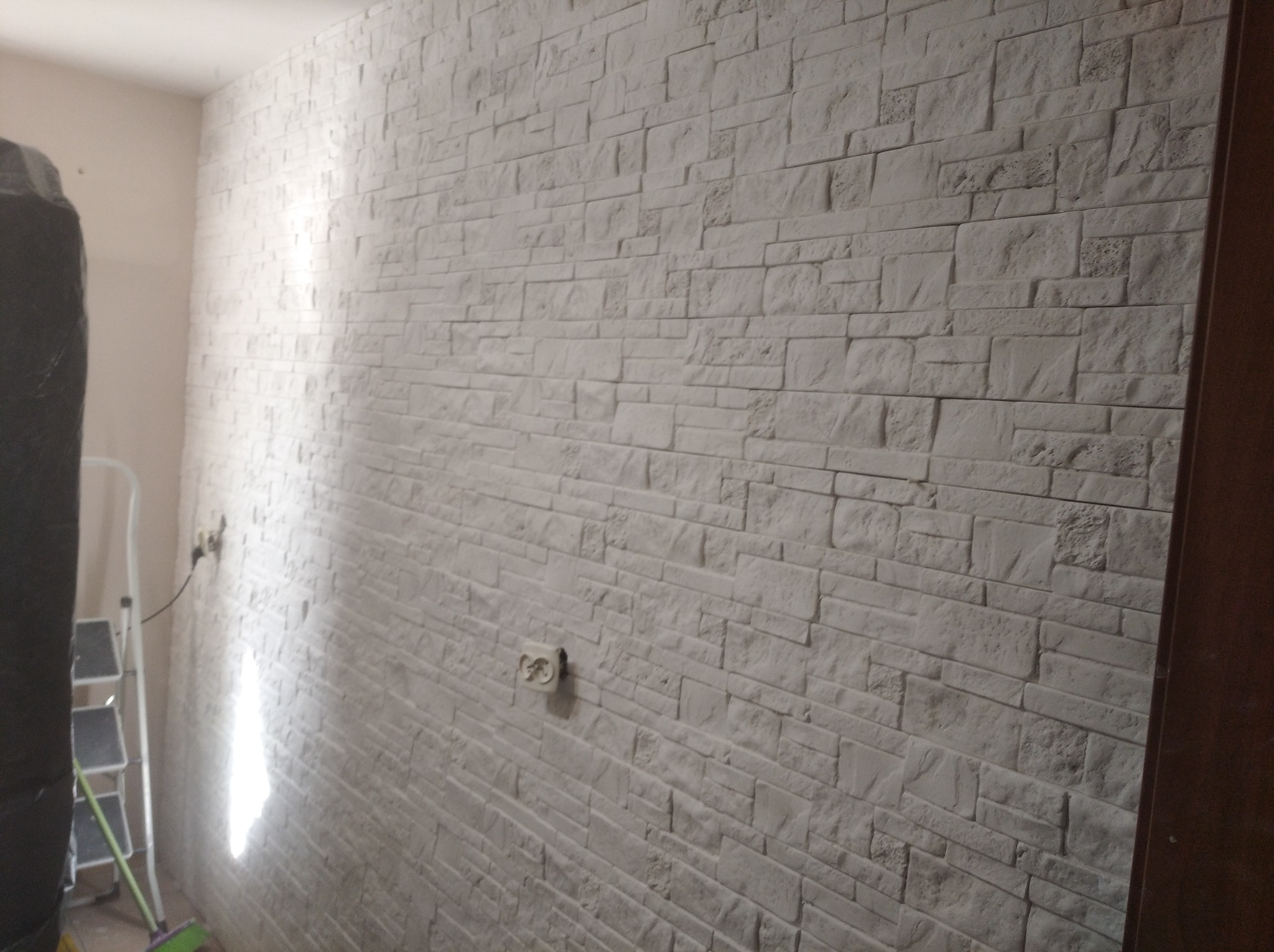 Wnętrze z wykończoną ścianą imitującą kamień, widoczna drabina malarska i gniazdko elektryczne.