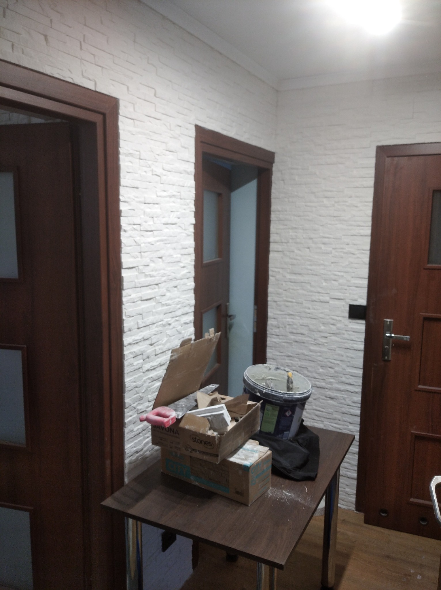 Wnętrze korytarza z trzema drewnianymi drzwiami i ścianą pokrytą dekoracyjną białą cegłą, na pierwszym planie stół z narzędziami i pojemnikiem z zaprawą.