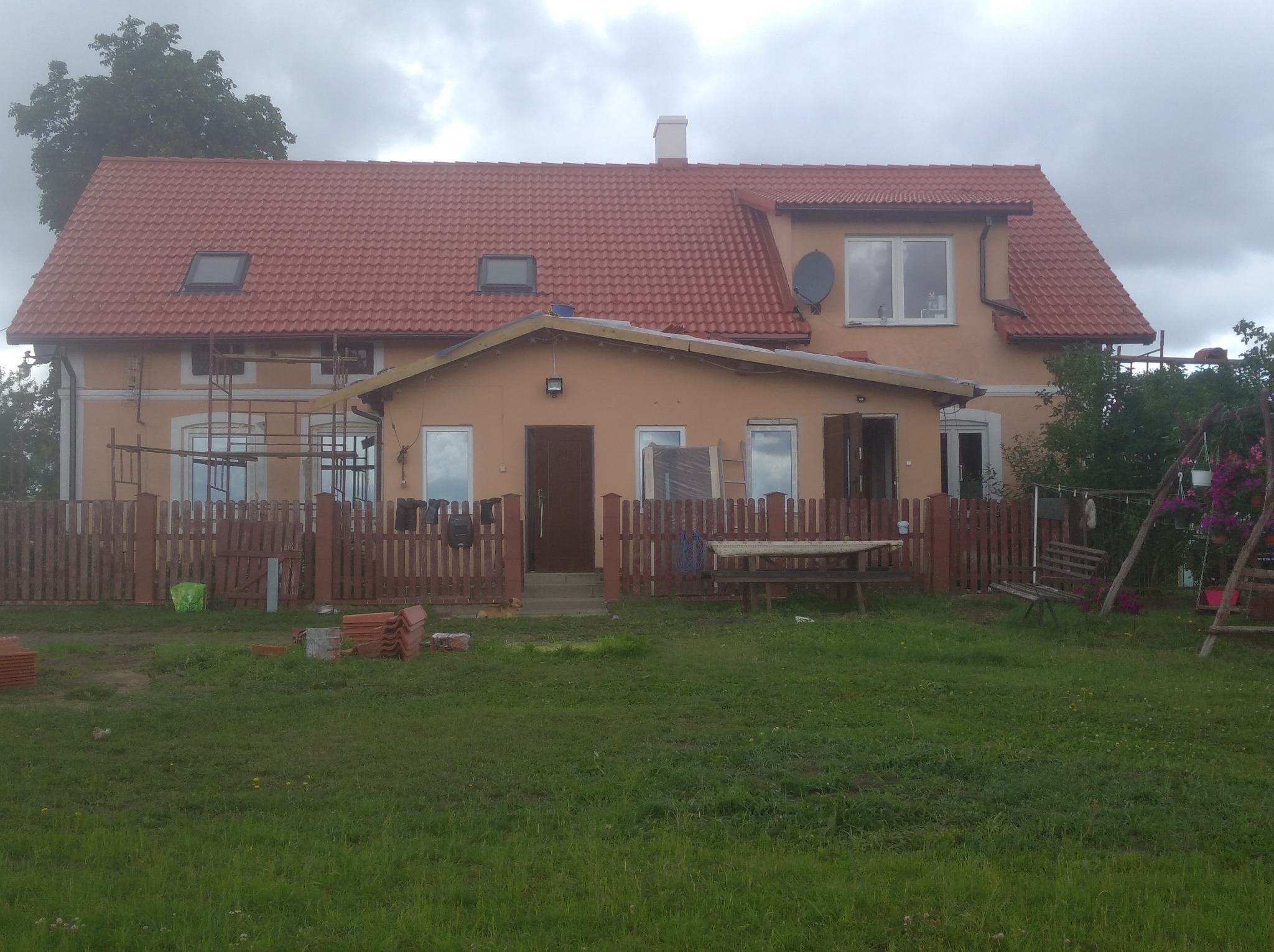 Dom jednorodzinny z nowym, czerwonym dachem z dachówki, widoczne rusztowania przy elewacji, dachówki na trawniku i drewniany płot.