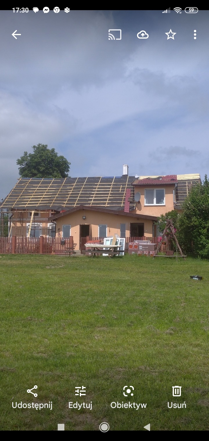 Dom w trakcie remontu dachu, widoczne krokwie i membrana dachowa, częściowo położona dachówka, okna i drzwi przygotowane do montażu na trawniku.