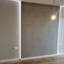 Ściana w pokoju po remoncie, z szarą dekoracyjną strukturą, oświetlona pionowymi panelami LED po bokach, widoczne gniazdka elektryczne i drewniana podłoga.