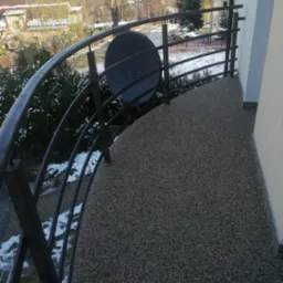 Balkon z szarą, kamyczkową posadzką żywiczną i metalową balustradą, widok na zimowy krajobraz z zasypanymi śniegiem drzewami.