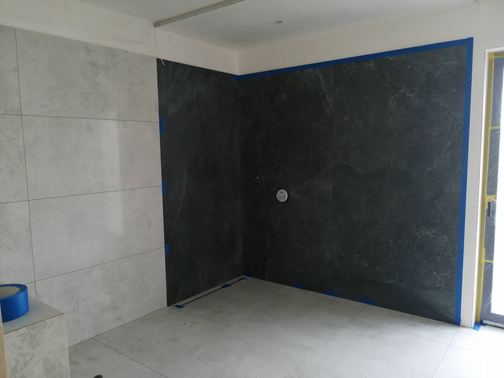 Łazienka w trakcie wykańczania: ułożone płytki w odcieniach szarości, zabezpieczone niebieską taśmą malarską, widoczny odpływ liniowy w podłodze i instalacja prysznicowa.