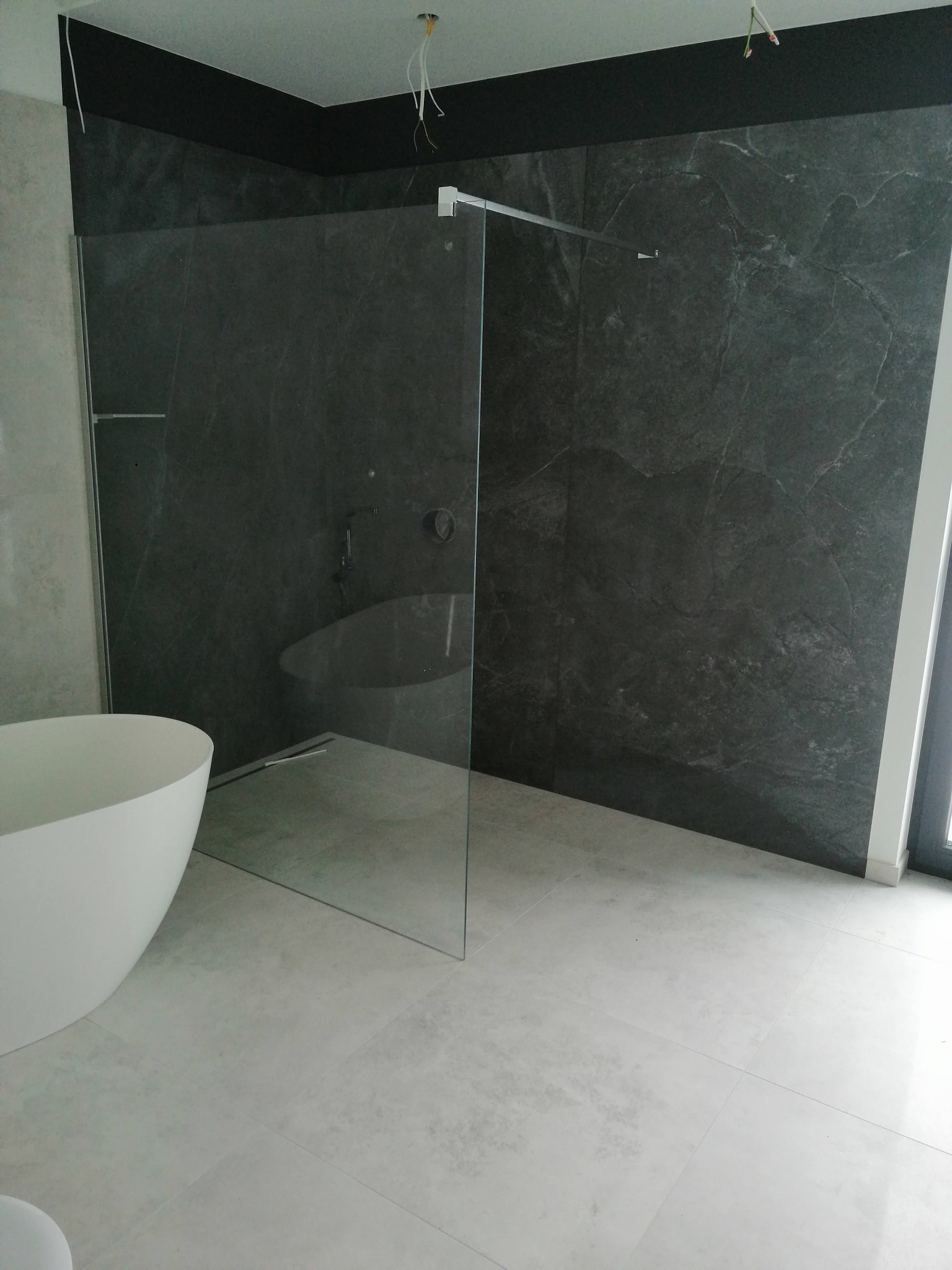 Nowoczesna łazienka z wolnostojącą białą wanną, szklaną kabiną prysznicową i ciemnoszarą ścianą z efektem kamienia.