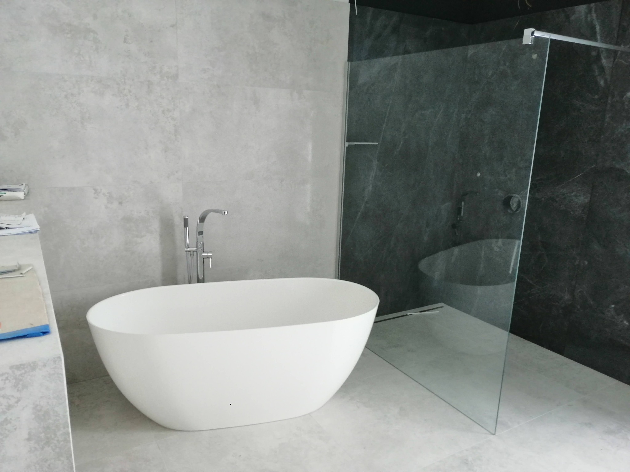 Nowoczesna łazienka z wolnostojącą białą wanną, chromowaną baterią, szarą marmurową ścianą i kabiną prysznicową ze szklaną ścianką.