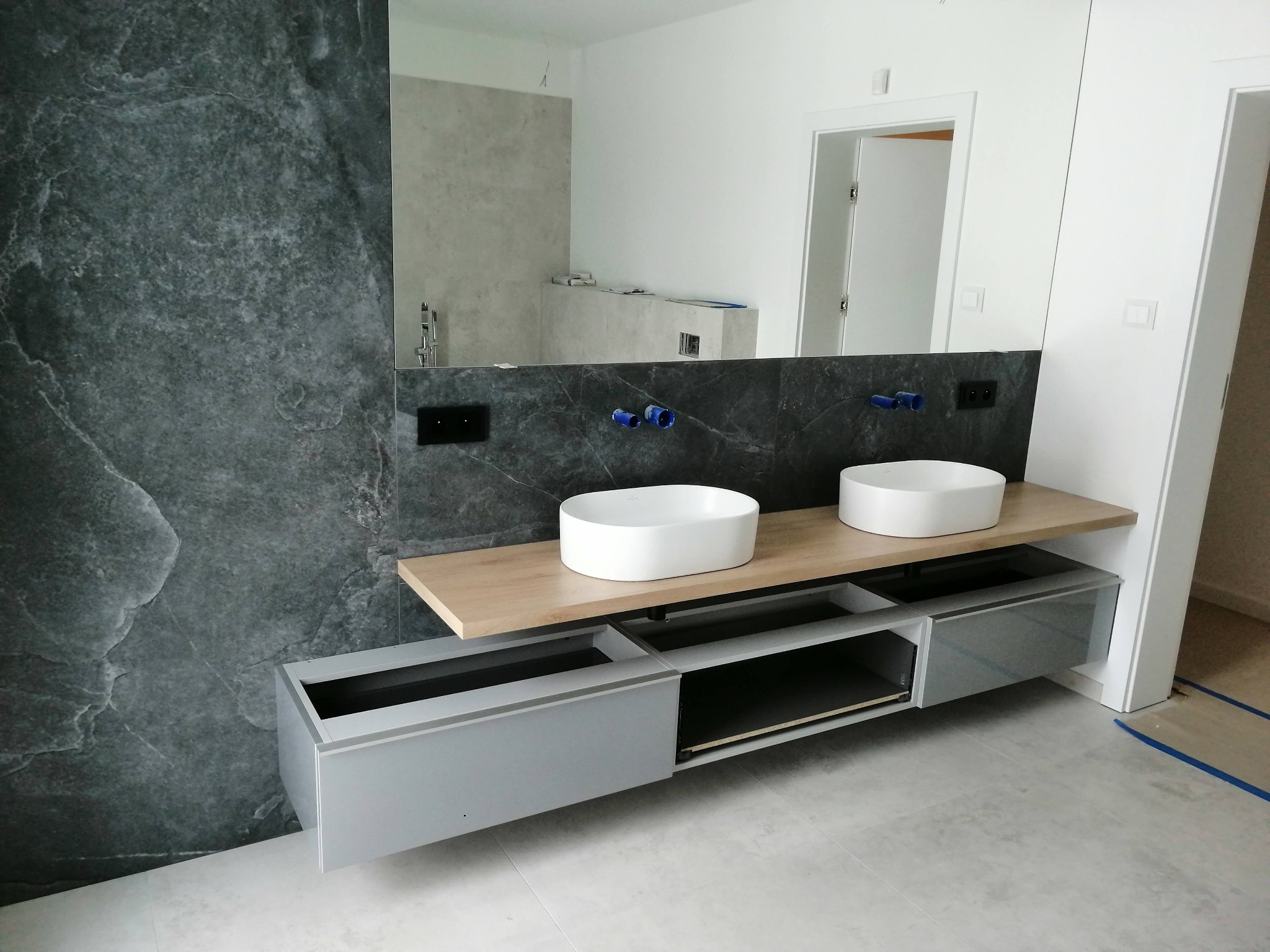 Nowoczesna łazienka z dwiema białymi umywalkami nablatowymi na drewnianym blacie, szara szafka pod umywalką z otwartymi półkami i szufladami, ściana wyłożona płytkami imitującymi ciemny marmur...