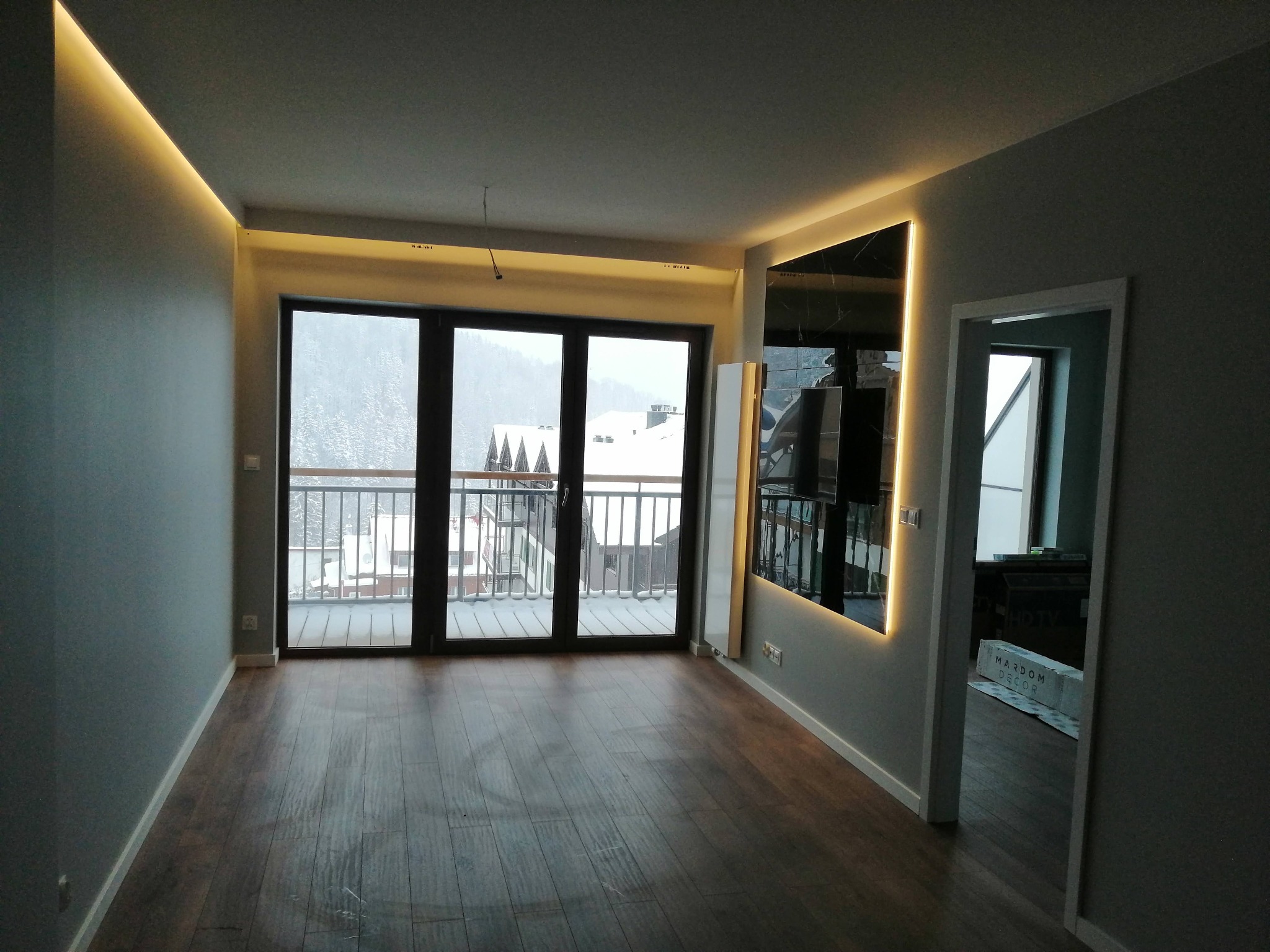 Wnętrze pokoju z drewnianą podłogą i szarymi ścianami, oświetlenie LED w suficie i wokół lustra, widok na zasypany śniegiem balkon i góry