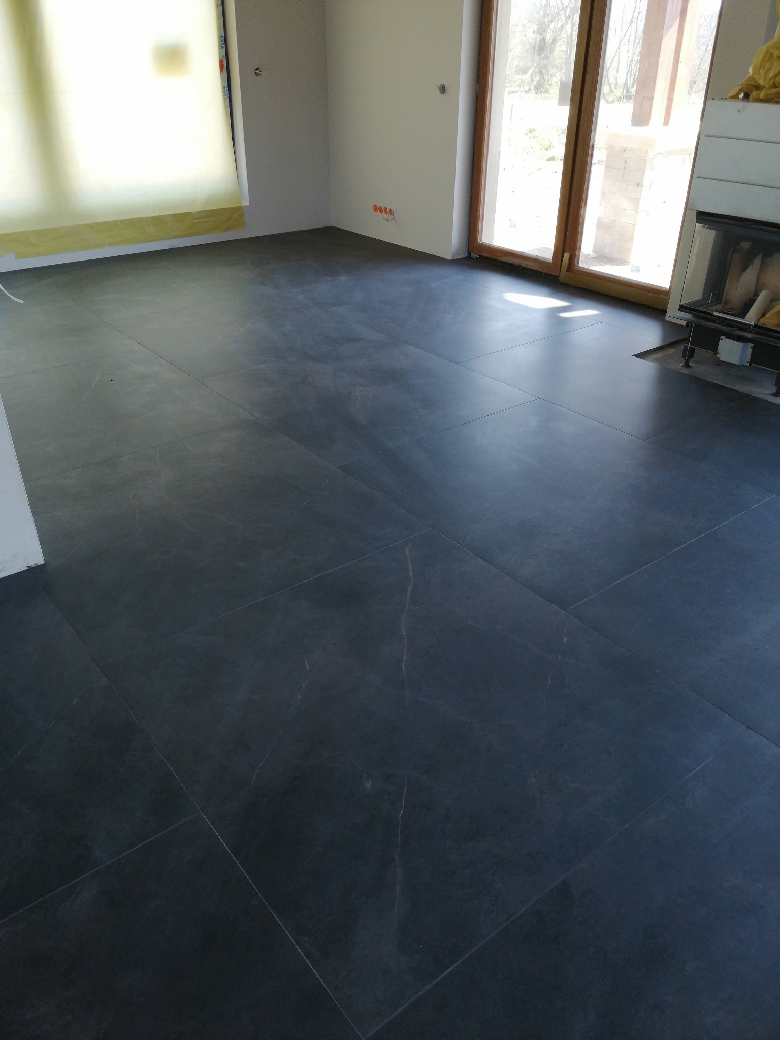 Nowoczesna, ciemnoszara posadzka z płytek imitujących kamień w jasnym, przestronnym wnętrzu z dużymi oknami i kominkiem.