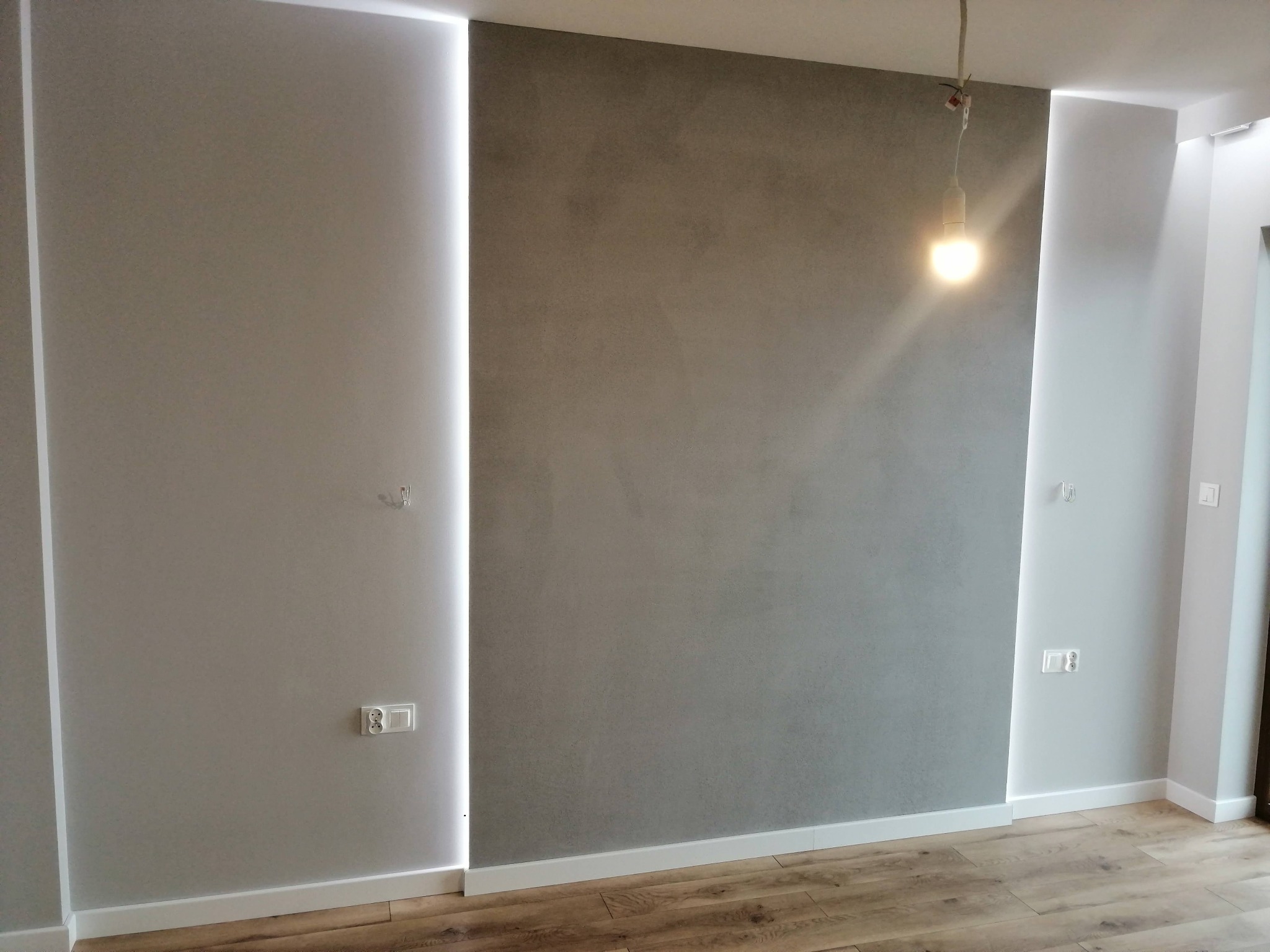 Ściana w pokoju po remoncie, z szarą dekoracyjną strukturą, oświetlona pionowymi panelami LED po bokach, widoczne gniazdka elektryczne i drewniana podłoga.