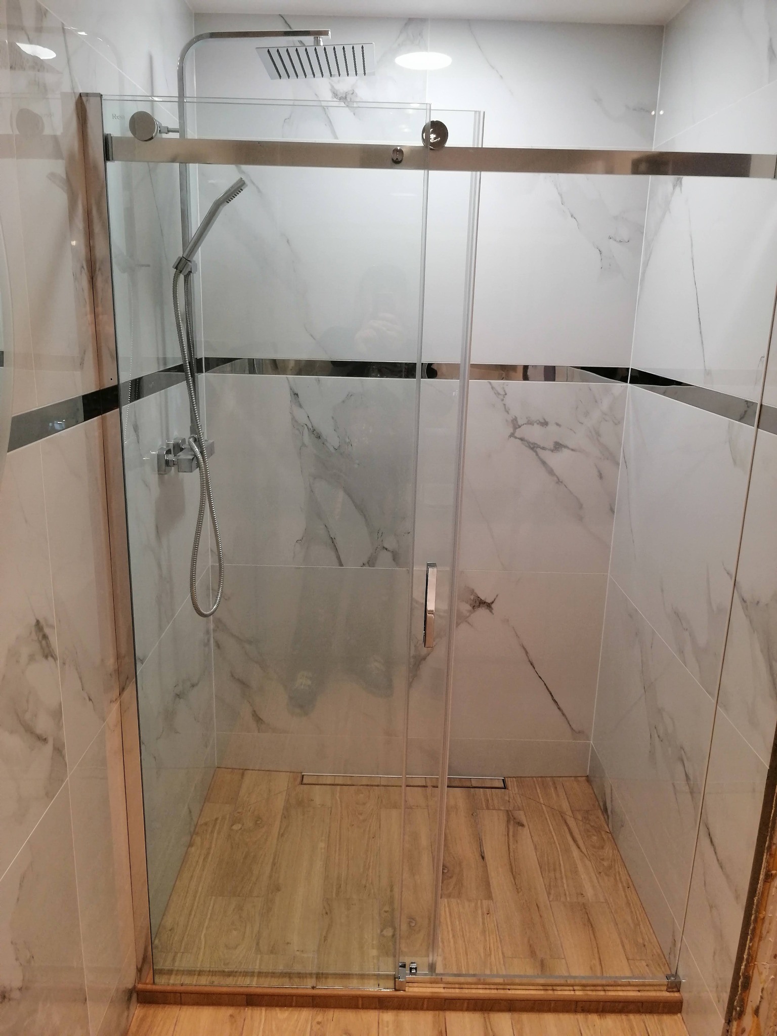 Nowoczesna kabina prysznicowa z przesuwnymi szklanymi drzwiami, wykończona płytkami imitującymi marmur i drewnopodobną podłogą z odpływem liniowym.