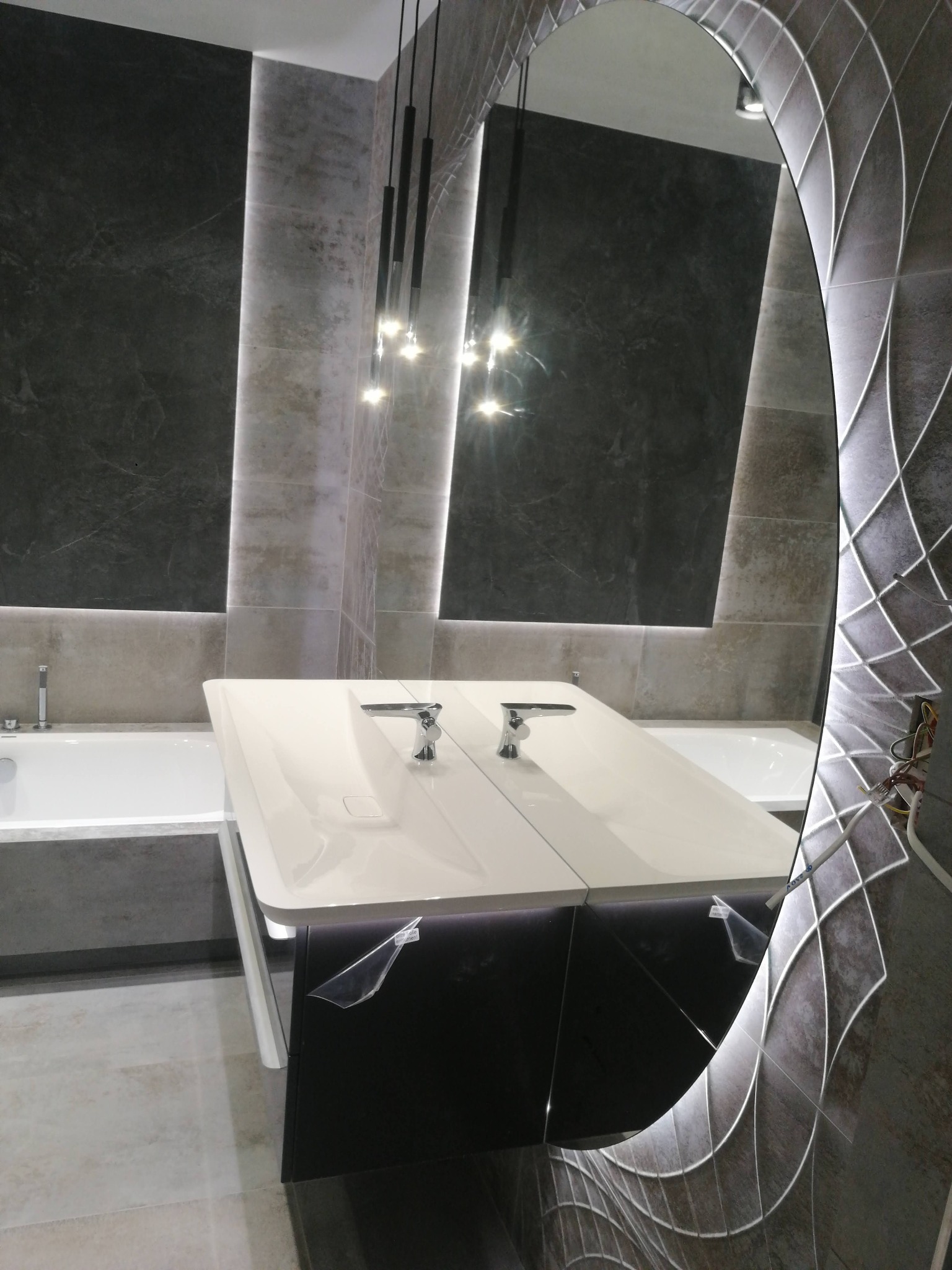 Nowoczesna łazienka z podwójną umywalką, oświetleniem LED i dekoracyjnym lustrem z podświetleniem. Na ścianach szare płytki w dwóch odcieniach, nad umywalką wiszące lampy. Widoczne zabezpieczenia...