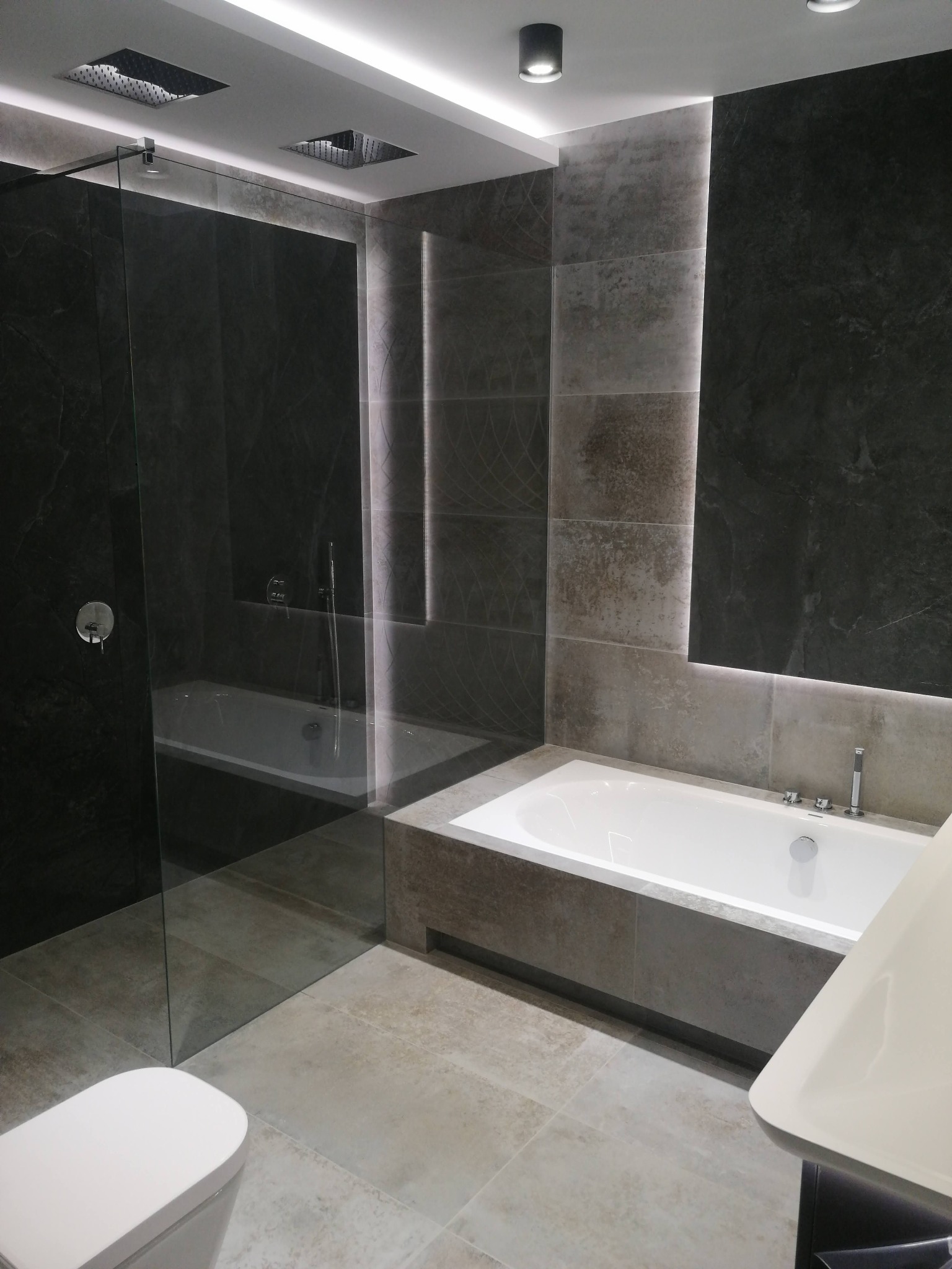 Nowoczesna łazienka z wanną, kabiną prysznicową typu walk-in, szarymi płytkami imitującymi kamień i podświetleniem LED wzdłuż ściany.