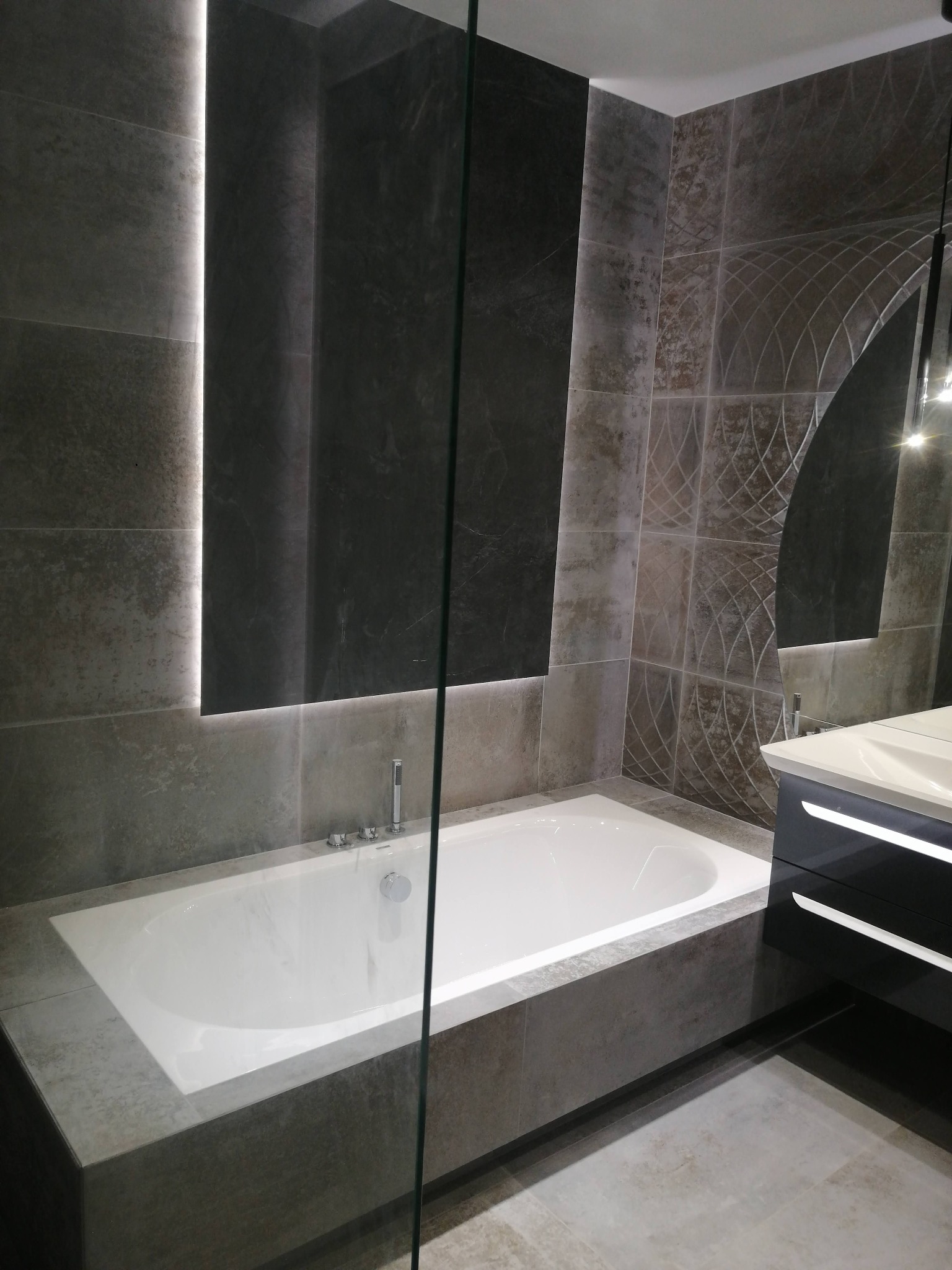 Nowoczesna łazienka z wanną obudowaną szarymi płytkami, podświetlonym panelem ściennym i umywalką z szafką w czarnym połysku.