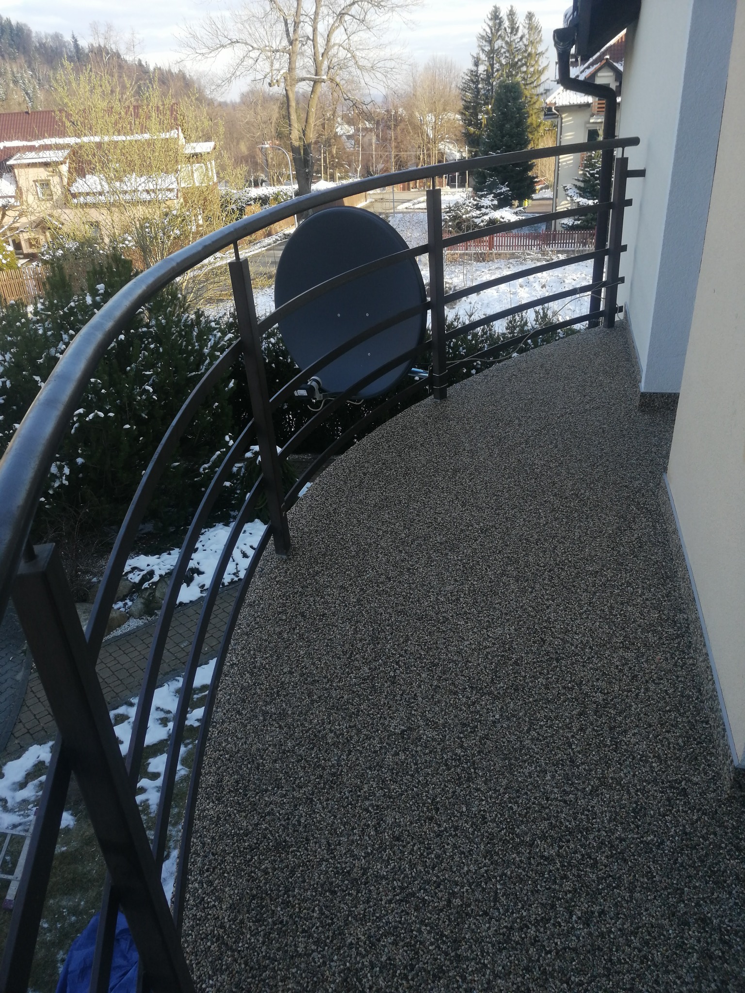 Balkon z szarą, kamyczkową posadzką żywiczną i metalową balustradą, widok na zimowy krajobraz z zasypanymi śniegiem drzewami.