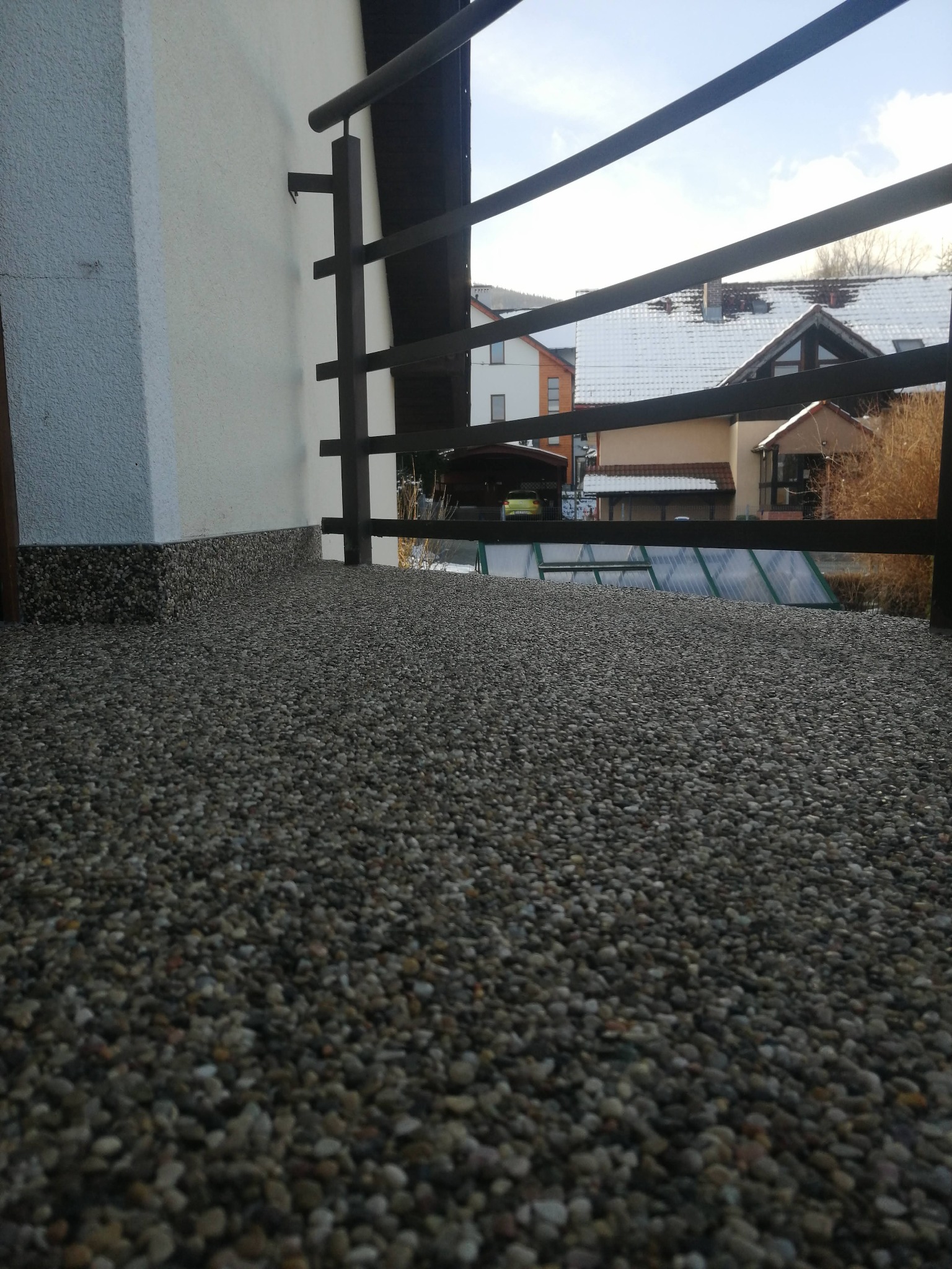 Balkon z wykończeniem posadzki z drobnych kamyczków, widok na zabudowę jednorodzinną zza czarnej, metalowej balustrady.