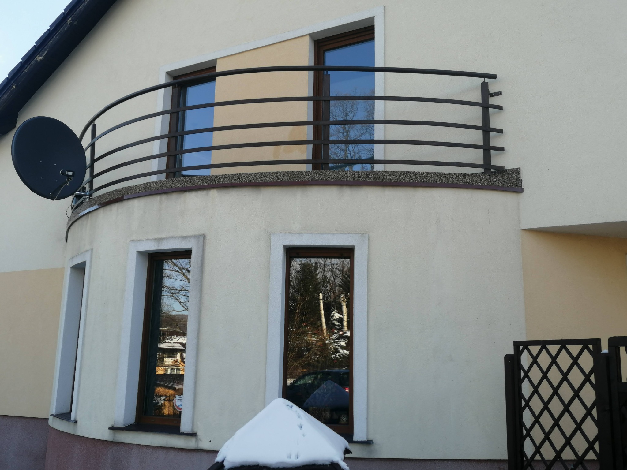 Jasna elewacja domu z zaokrąglonym balkonem z czarną, metalową balustradą, na której zamontowana jest antena satelitarna; widoczne okna z odbiciami zimowego krajobrazu.