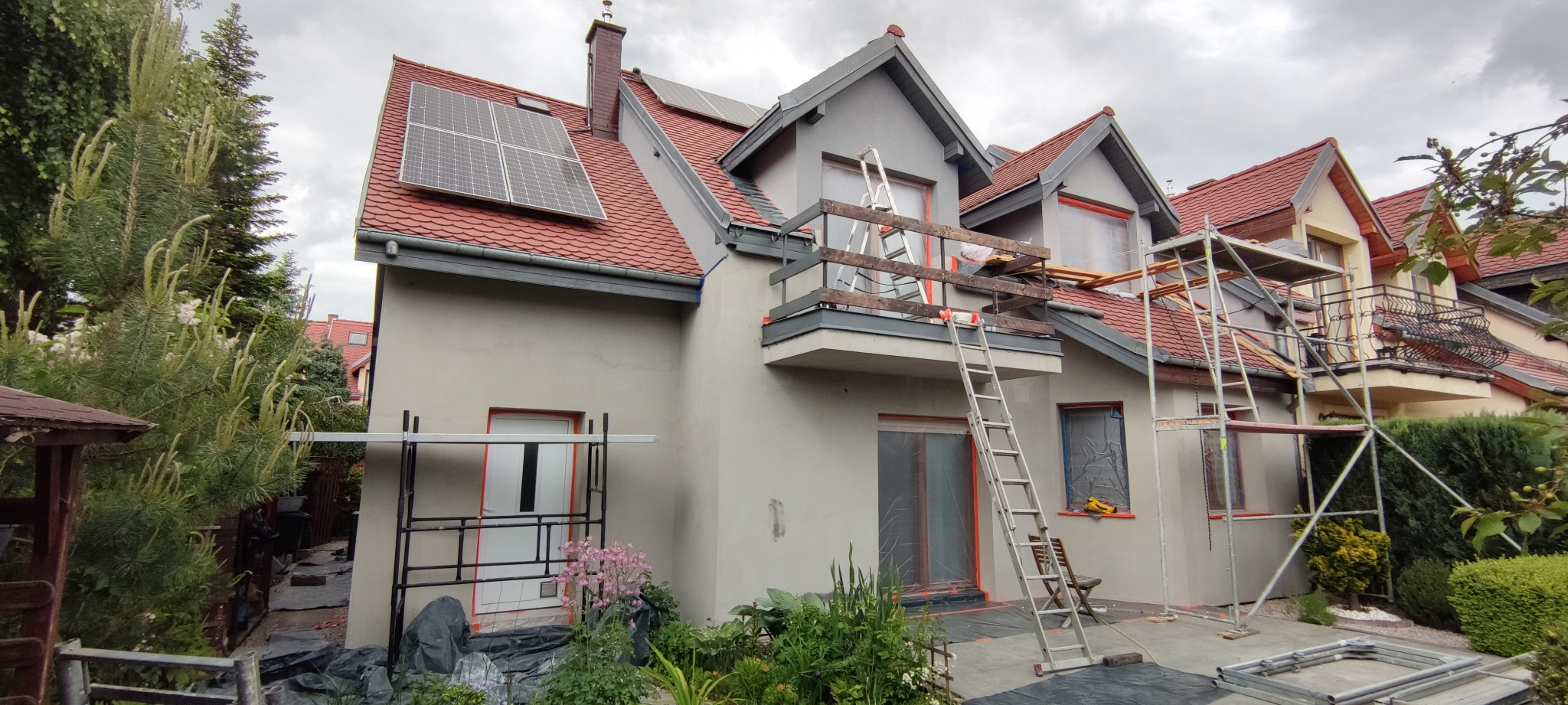 Dom jednorodzinny w trakcie prac elewacyjnych: elewacja pokryta szarym tynkiem, okna zabezpieczone folią, rusztowania ustawione przy ścianach, drabina oparta o balkon, panele słoneczne na dachu.
