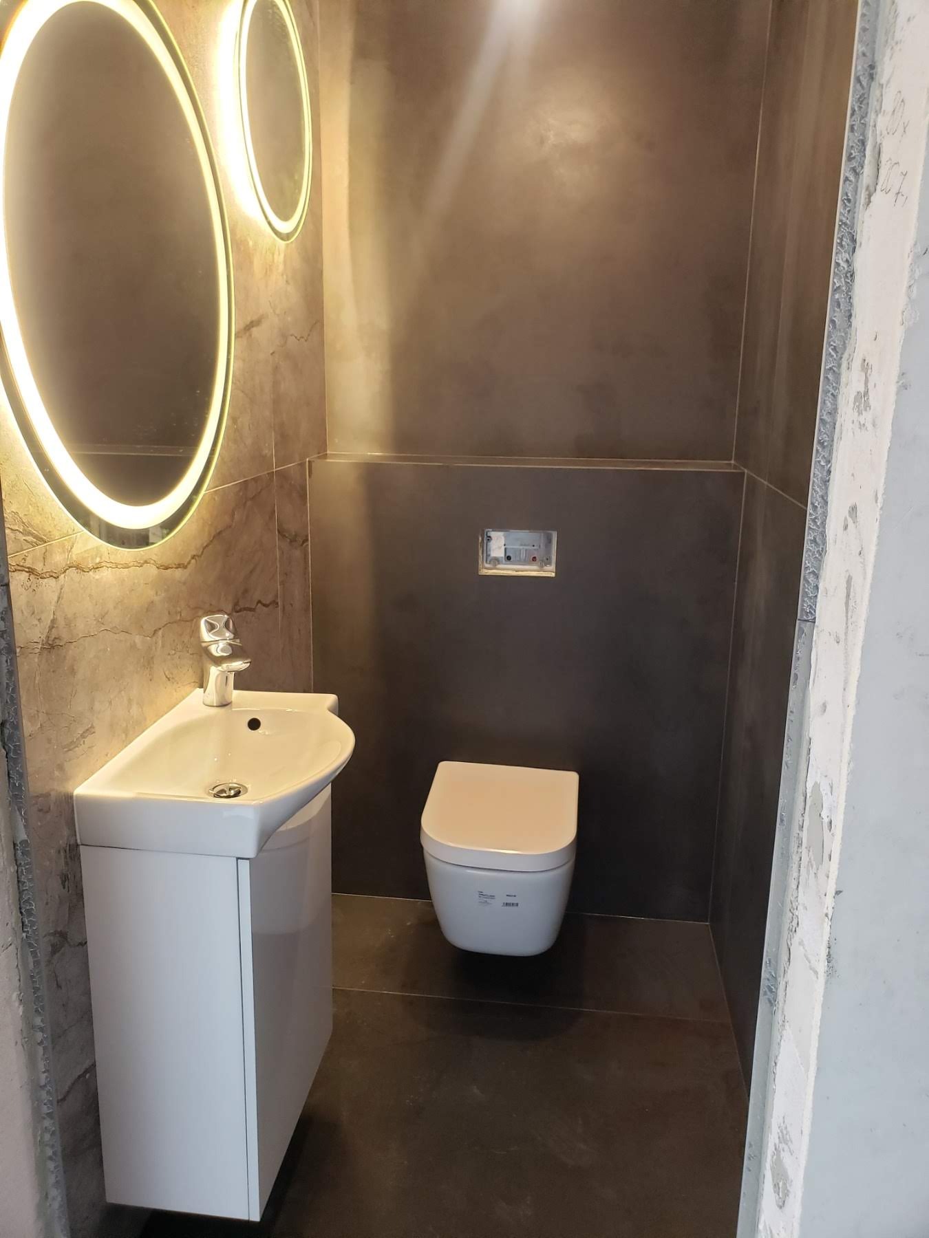 Nowoczesna, minimalistyczna łazienka z umywalką na szafce, podświetlanymi lustrami i podwieszaną toaletą na tle szarych ścian z efektem betonu.