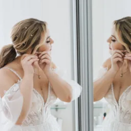 Panna młoda w koronkowej sukni, z włosami upiętymi perłami, zakłada kolczyki przed lustrem, przygotowując się do ślubu.