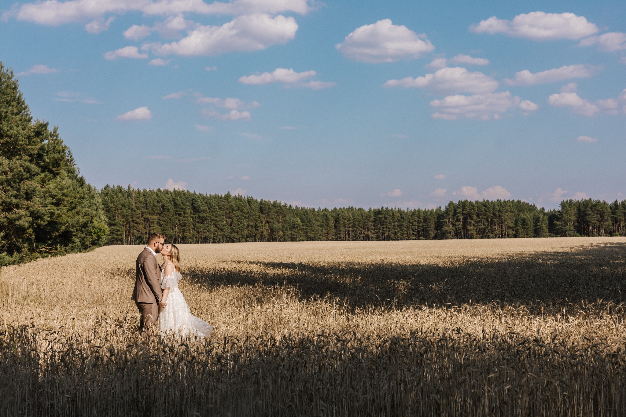 Para młoda w objęciach na tle pola zboża i lasu w słoneczny dzień, panna młoda w zwiewnej sukni z koronkowymi rękawami, pan młody w brązowym garniturze.