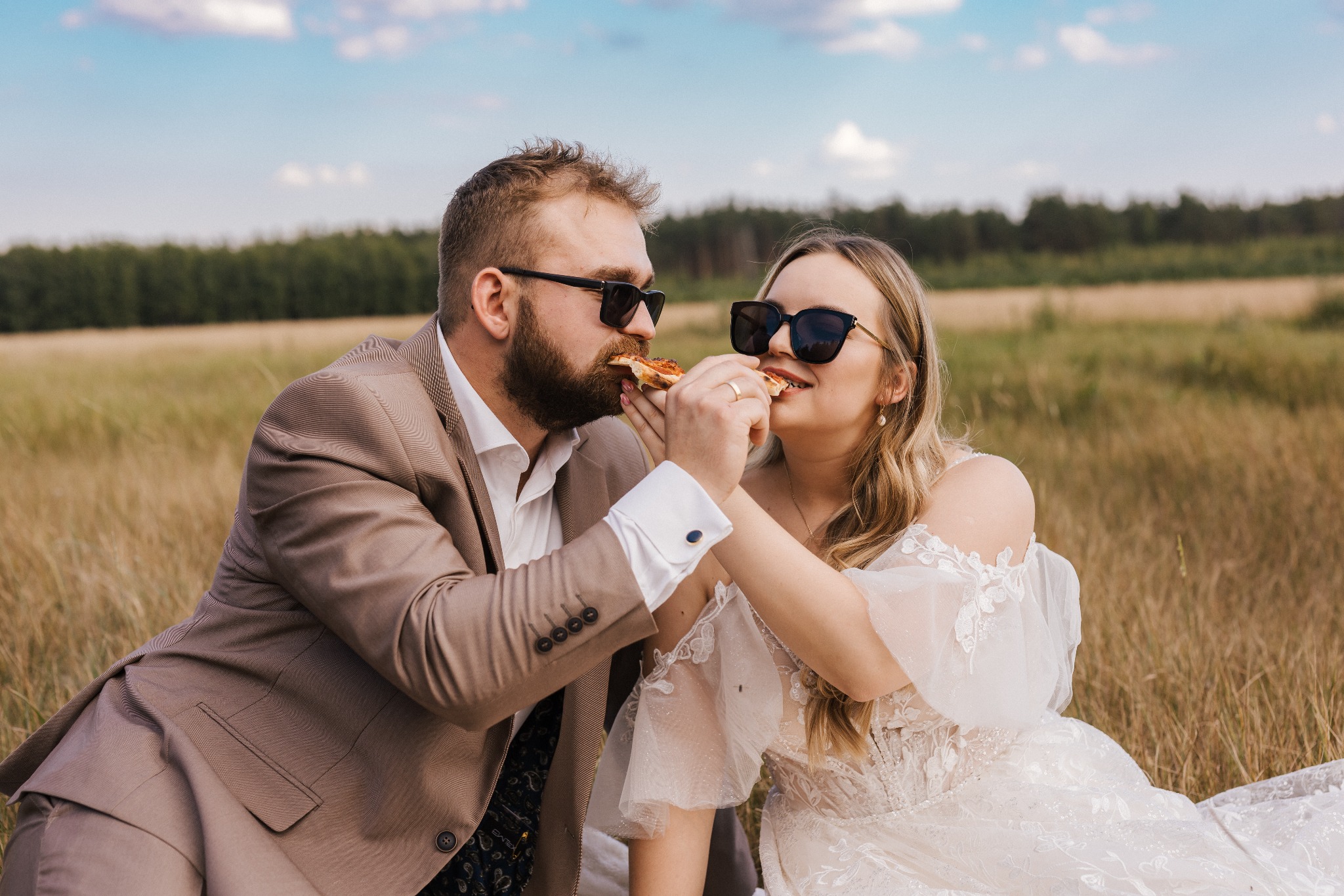 Para młoda w okularach przeciwsłonecznych dzieli się kawałkiem pizzy na łonie natury, panna młoda w koronkowej sukni, pan młody w brązowym garniturze.