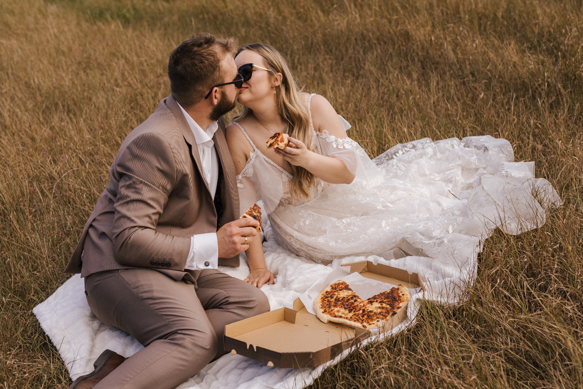 Para młoda w trawie, w sukni ślubnej i garniturze, jedząca pizzę na pikniku, z bliska, w okularach przeciwsłonecznych.