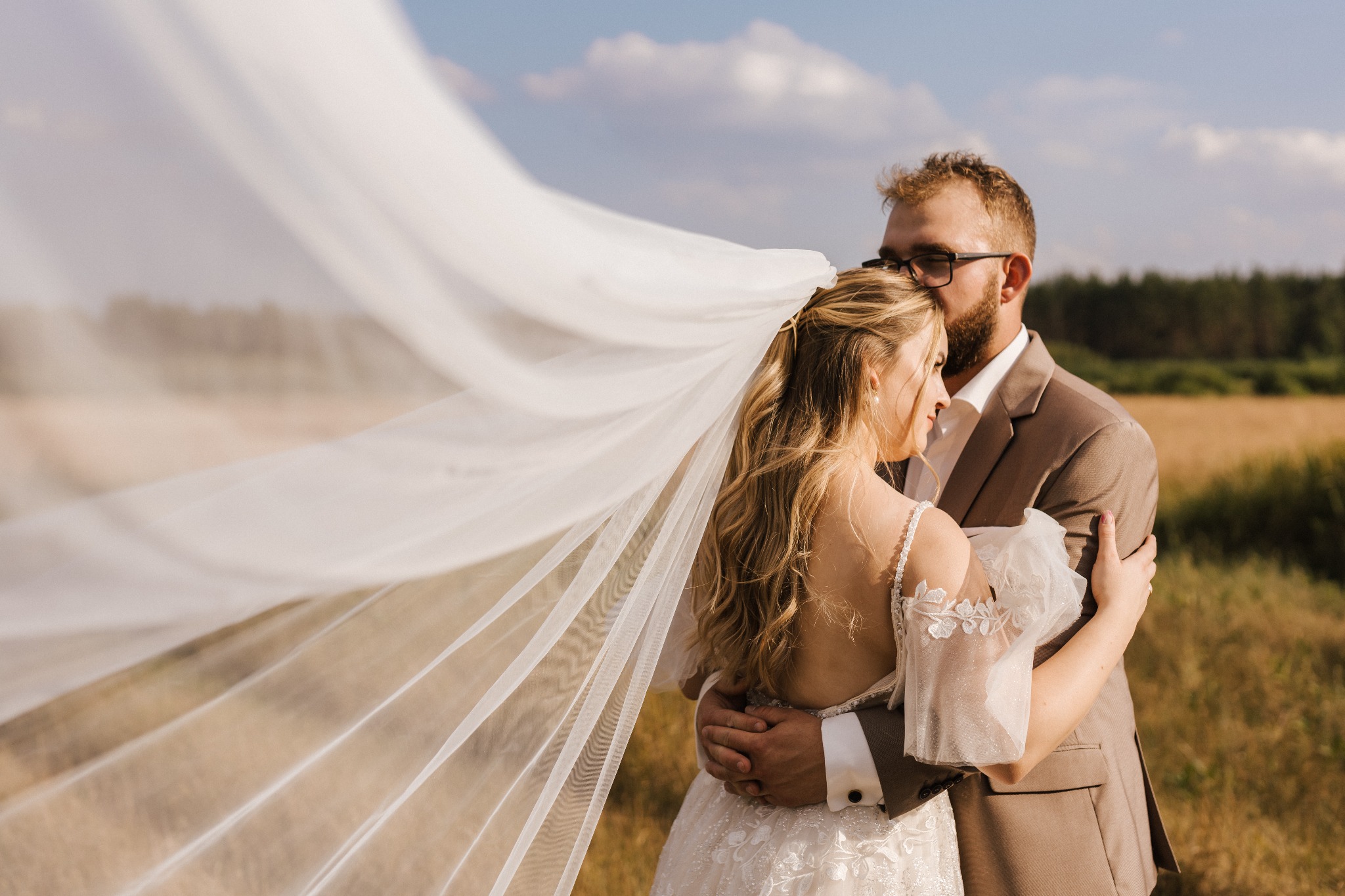 Para młoda w objęciach na tle pola, welon powiewa na wietrze, delikatne światło słoneczne.