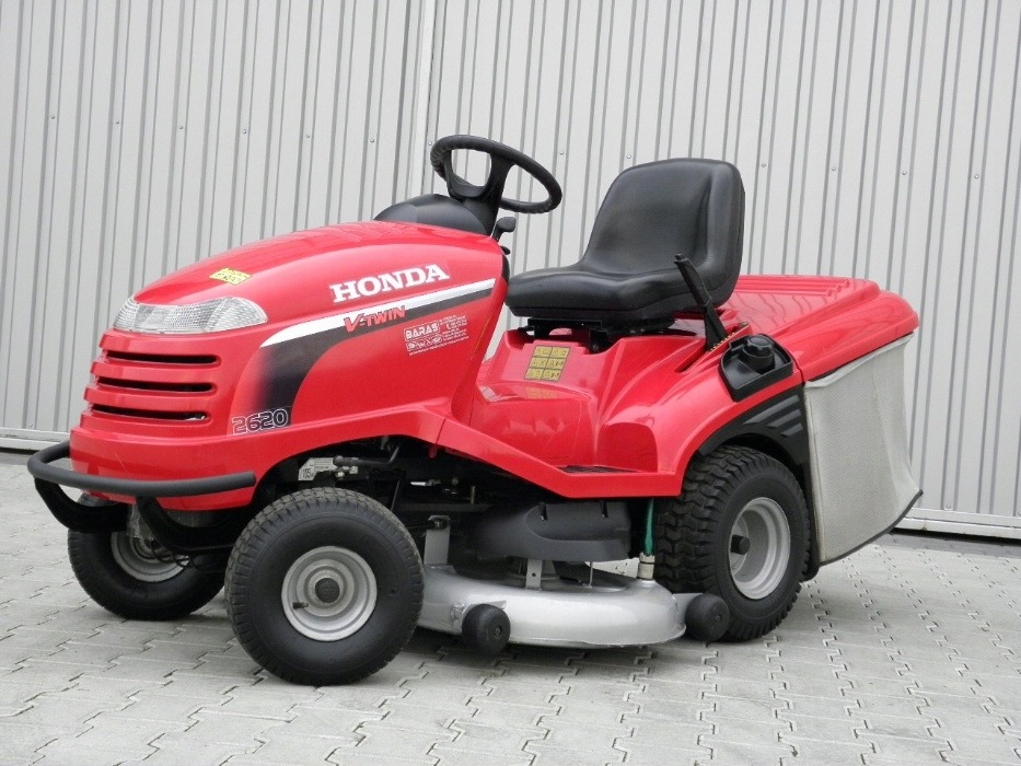 Czerwona kosiarka traktorek Honda 2620 V-Twin z czarnym siedzeniem i koszem na trawę, stojąca na szarej kostce brukowej przed szarą ścianą z blachy trapezowej.