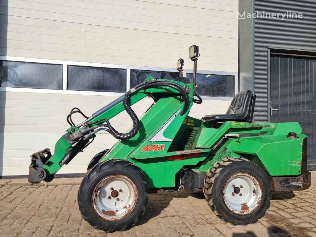 Zielona miniładowarka Avant z łyżką, widok z boku na tle białej ściany budynku z oknami i szarej ściany blaszanej z drzwiami.