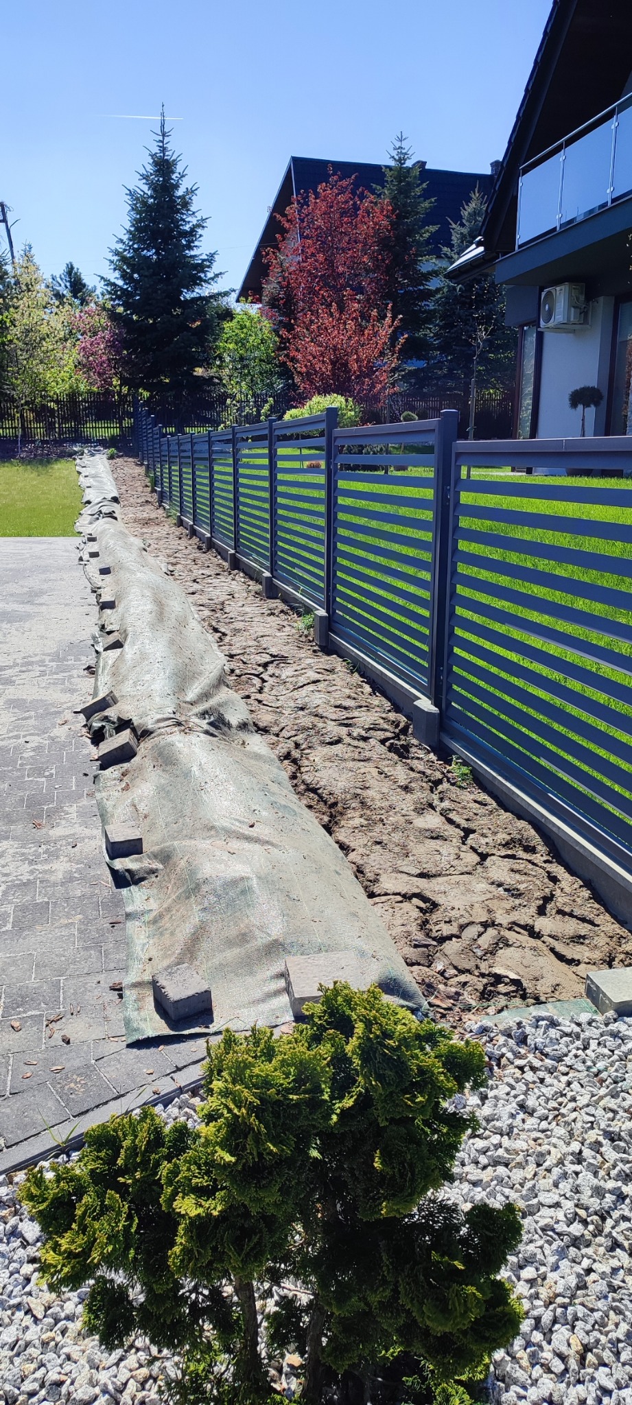 Montaż nowoczesnego ogrodzenia panelowego, przygotowanie terenu pod trawnik, widoczna warstwa ziemi i zabezpieczająca mata, w tle dom z balkonem i bujna roślinność.