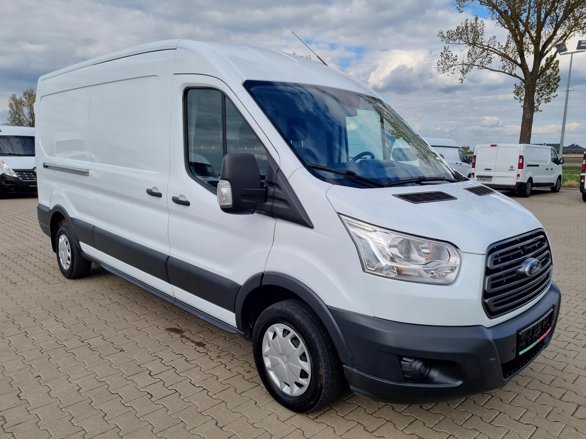 Biały Ford Transit, widok z przodu i boku, zaparkowany na kostce brukowej z innymi pojazdami w tle, lekko pochmurne niebo.