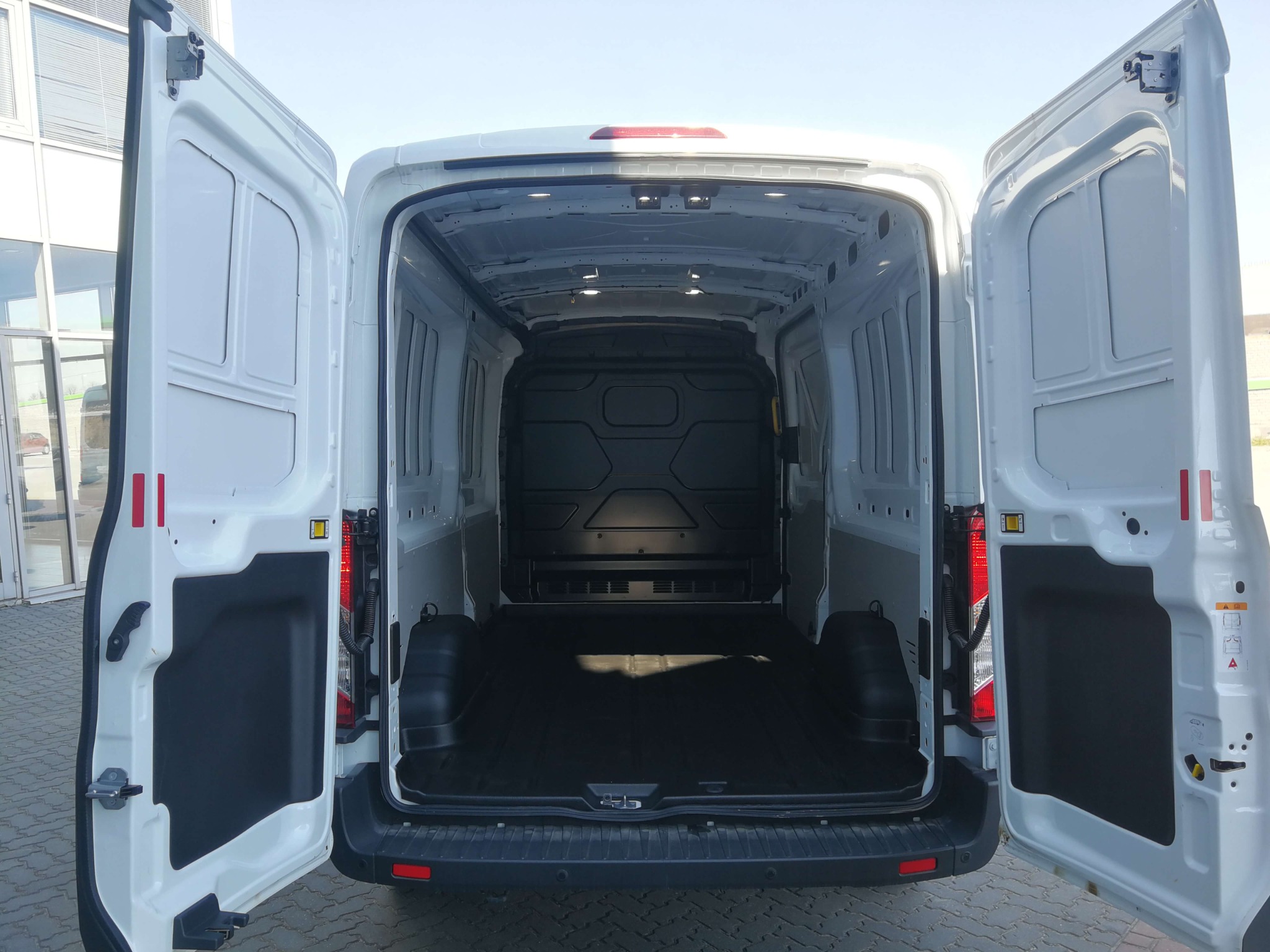 Wnętrze paki białego busa z otwartymi drzwiami, widok od tyłu na pustą, czystą przestrzeń ładunkową, z czarną gumową podłogą i panelami ochronnymi na ścianach.