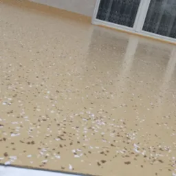 Beżowa powierzchnia balkonu z żywicy z drobnymi, białymi i brązowymi elementami dekoracyjnymi, widok z góry, okno z firanką w tle.