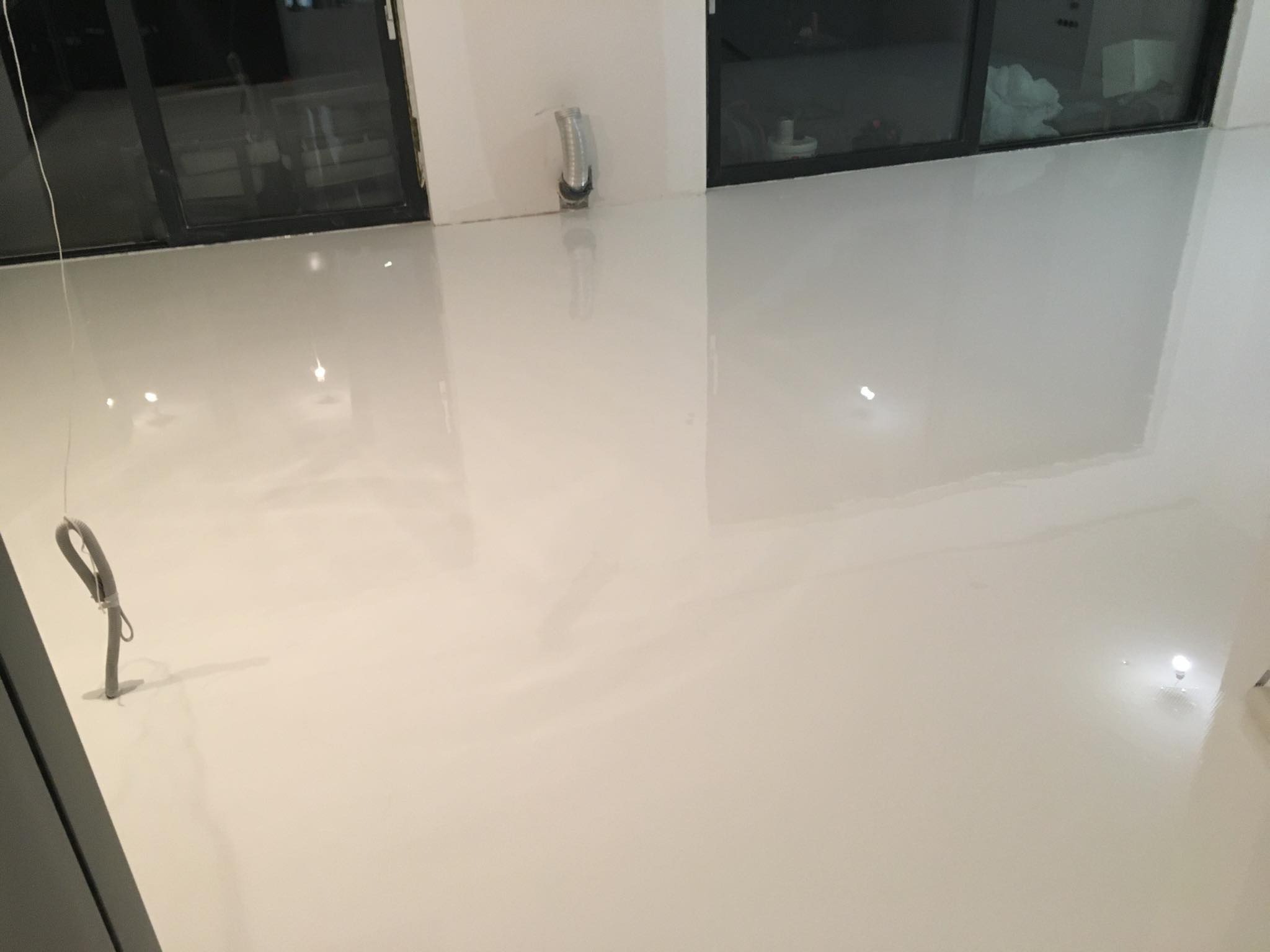 Biała, błyszcząca podłoga w jasnym pomieszczeniu z dużymi oknami, widoczne odbicia światła, przy ścianie wystająca instalacja elektryczna.