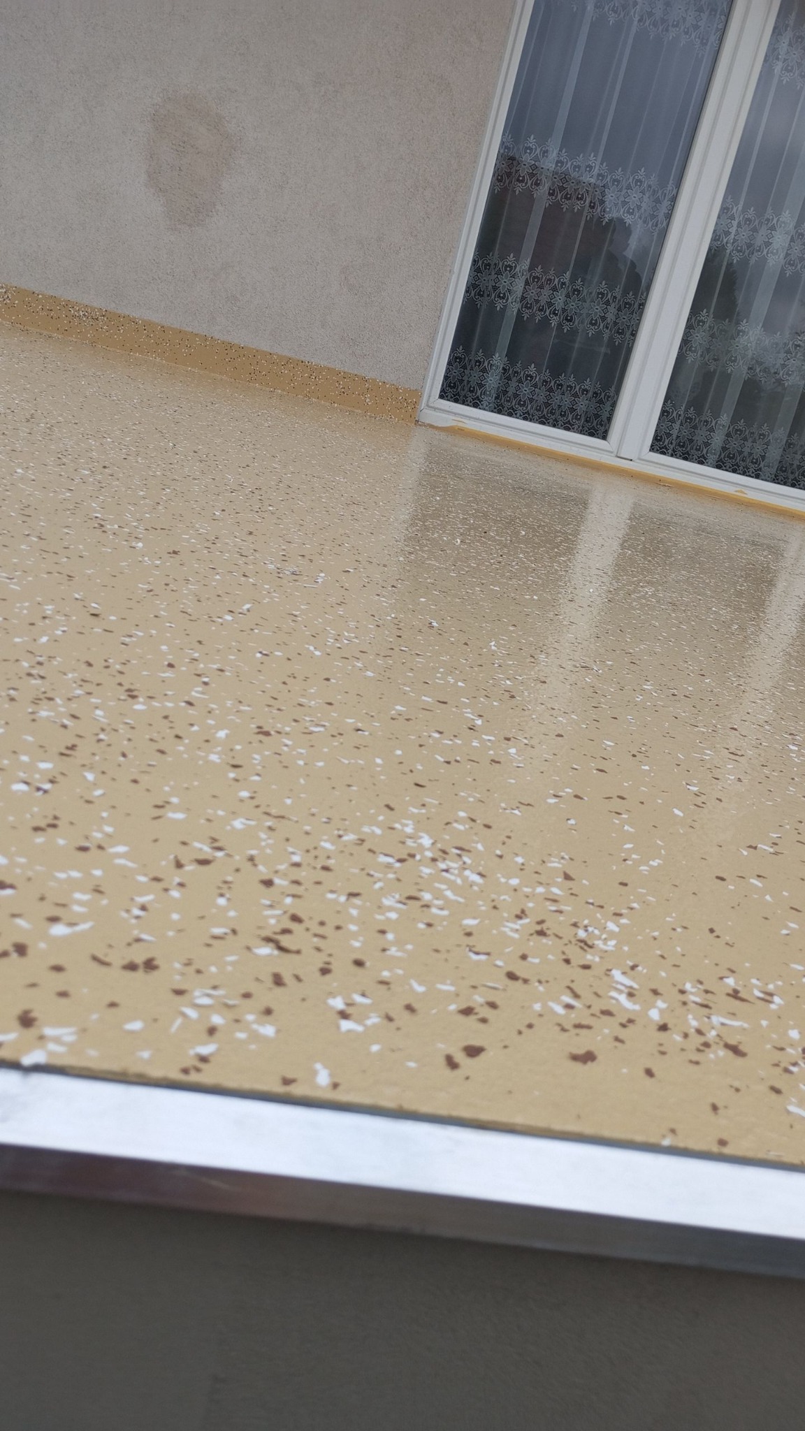 Beżowa powierzchnia balkonu z żywicy z drobnymi, białymi i brązowymi elementami dekoracyjnymi, widok z góry, okno z firanką w tle.