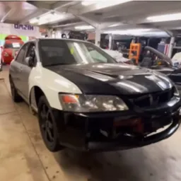 Mitsubishi Lancer Evolution w warsztacie, z elementami karoserii w różnych kolorach (srebrny, biały, czarny), sugerującymi naprawę lub modyfikację.