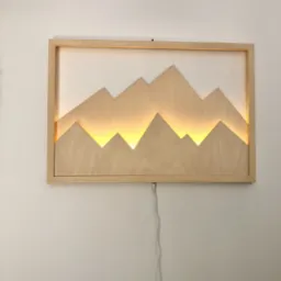 Drewniana, podświetlana dekoracja ścienna w kształcie gór w jasnej ramie, zawieszona na białej ścianie, z widocznym kablem zasilającym.